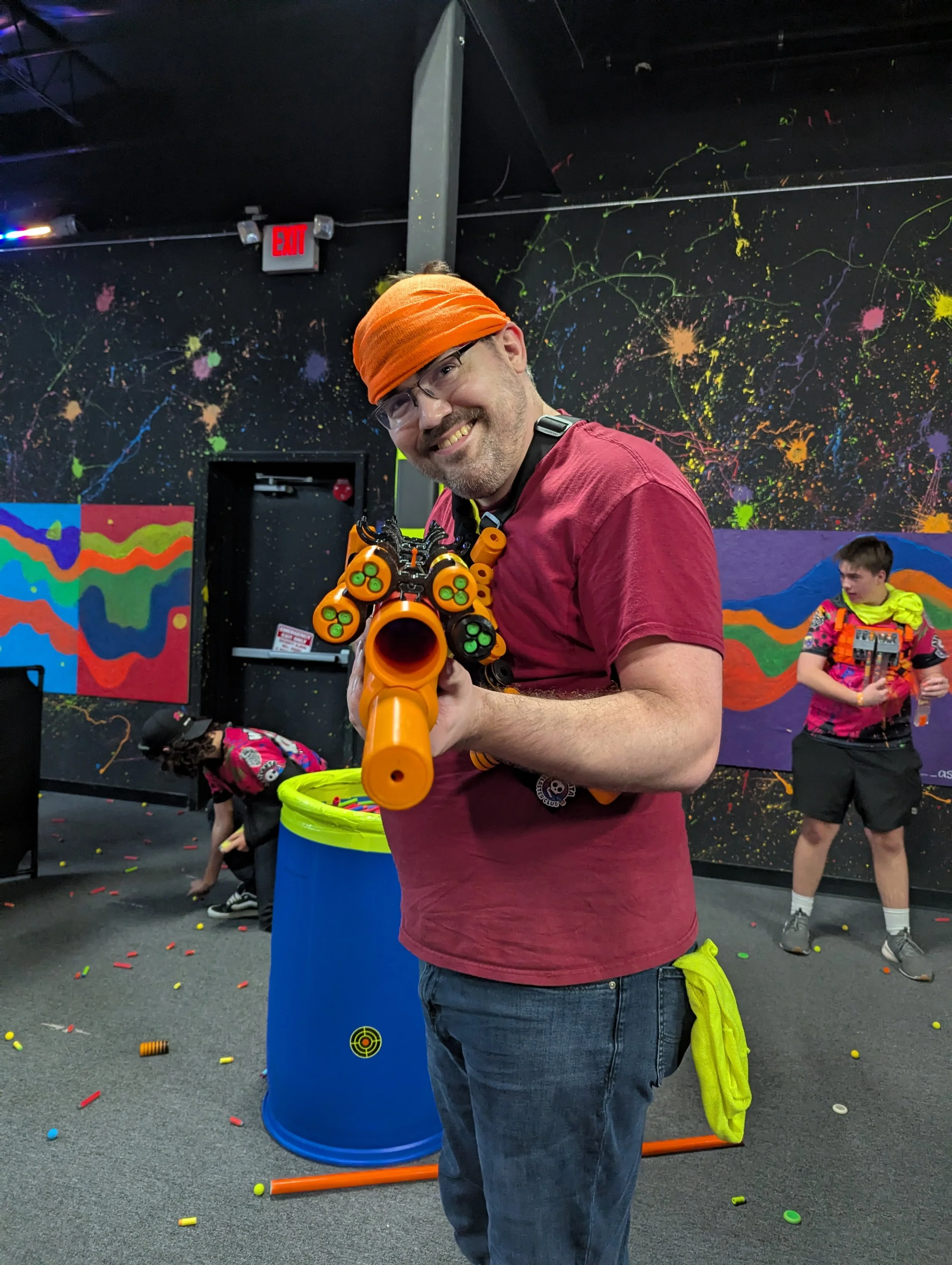 A man smiling and holding a toy gun-like object in an indoor play area with a black wall decorated with colorful paint splatters. A young boy in colorful clothing stands in the background among scattered toys and cones, with a blue barrel and orange 