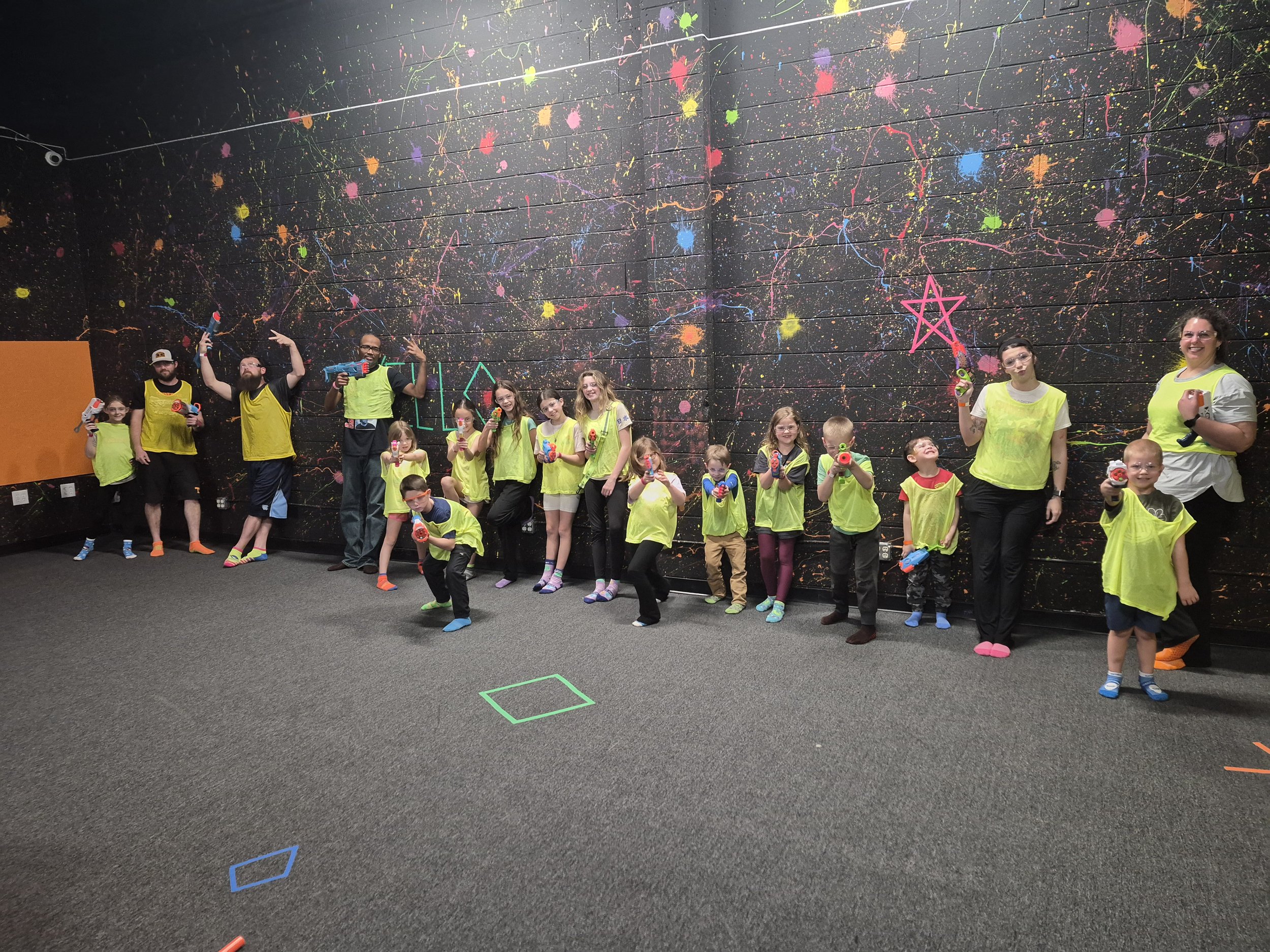Children and instructors in neon yellow and gray shirts at an indoor nerf battle or game facility, holding toy Nerf blasters. The background features a black wall with colorful splatter paint and neon drawings, while the group poses happily.