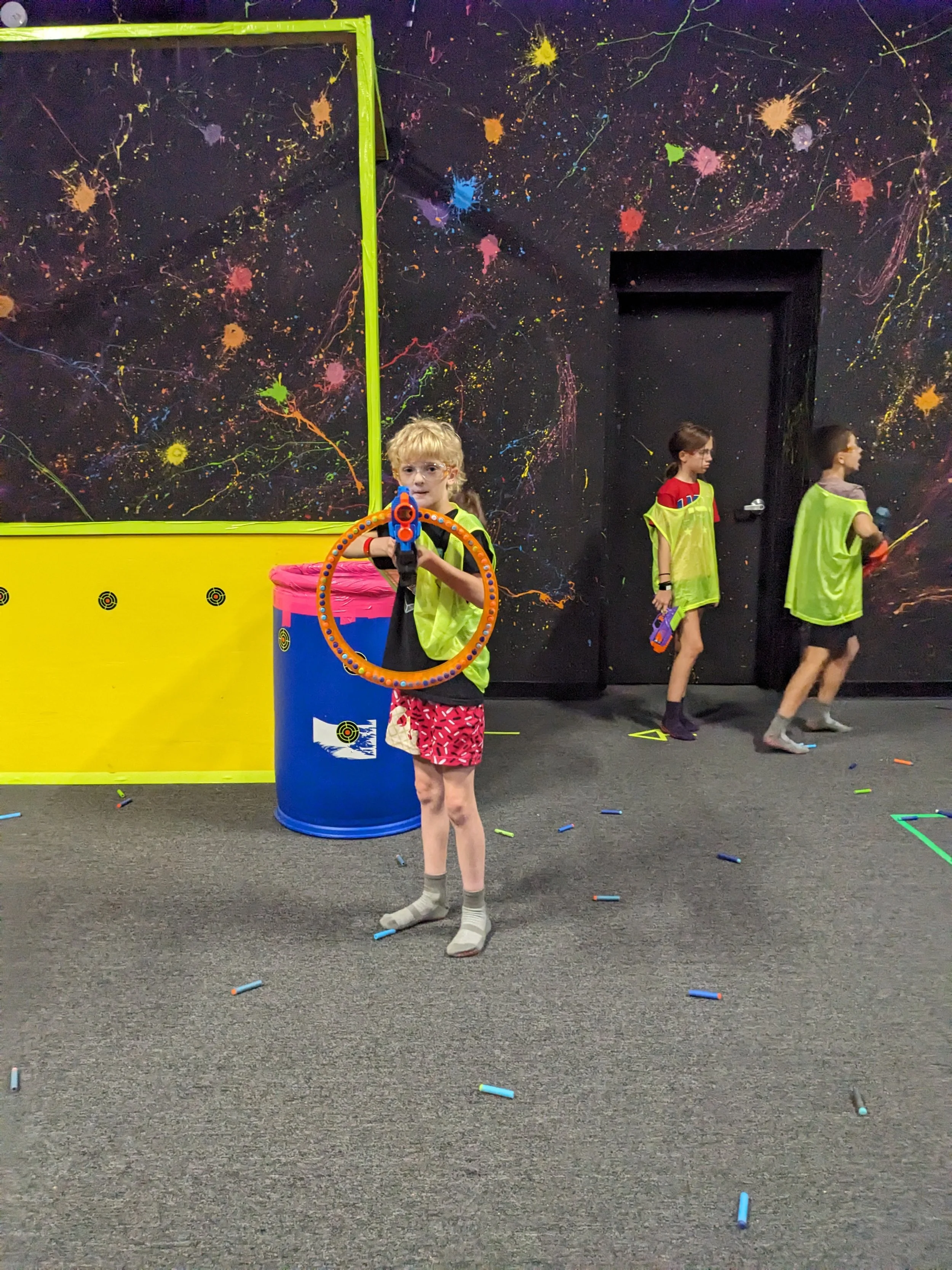 Children playing laser tag equipment in an indoor arcade with black walls decorated with colorful neon splatters.