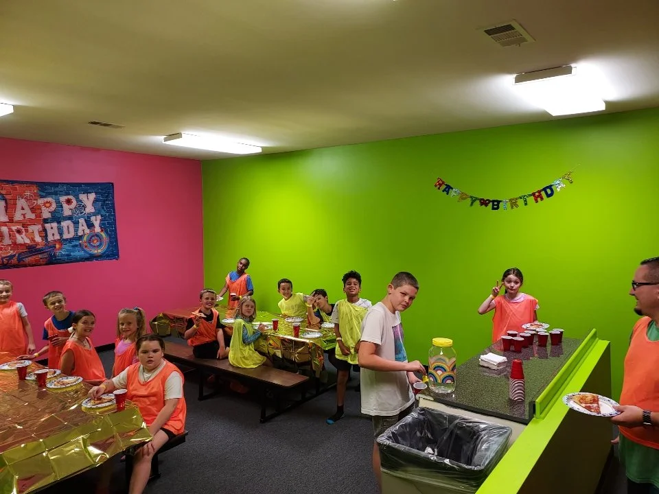 Children celebrating a birthday at a party with colorful walls, a 'Happy Birthday' banner, and party decorations.
