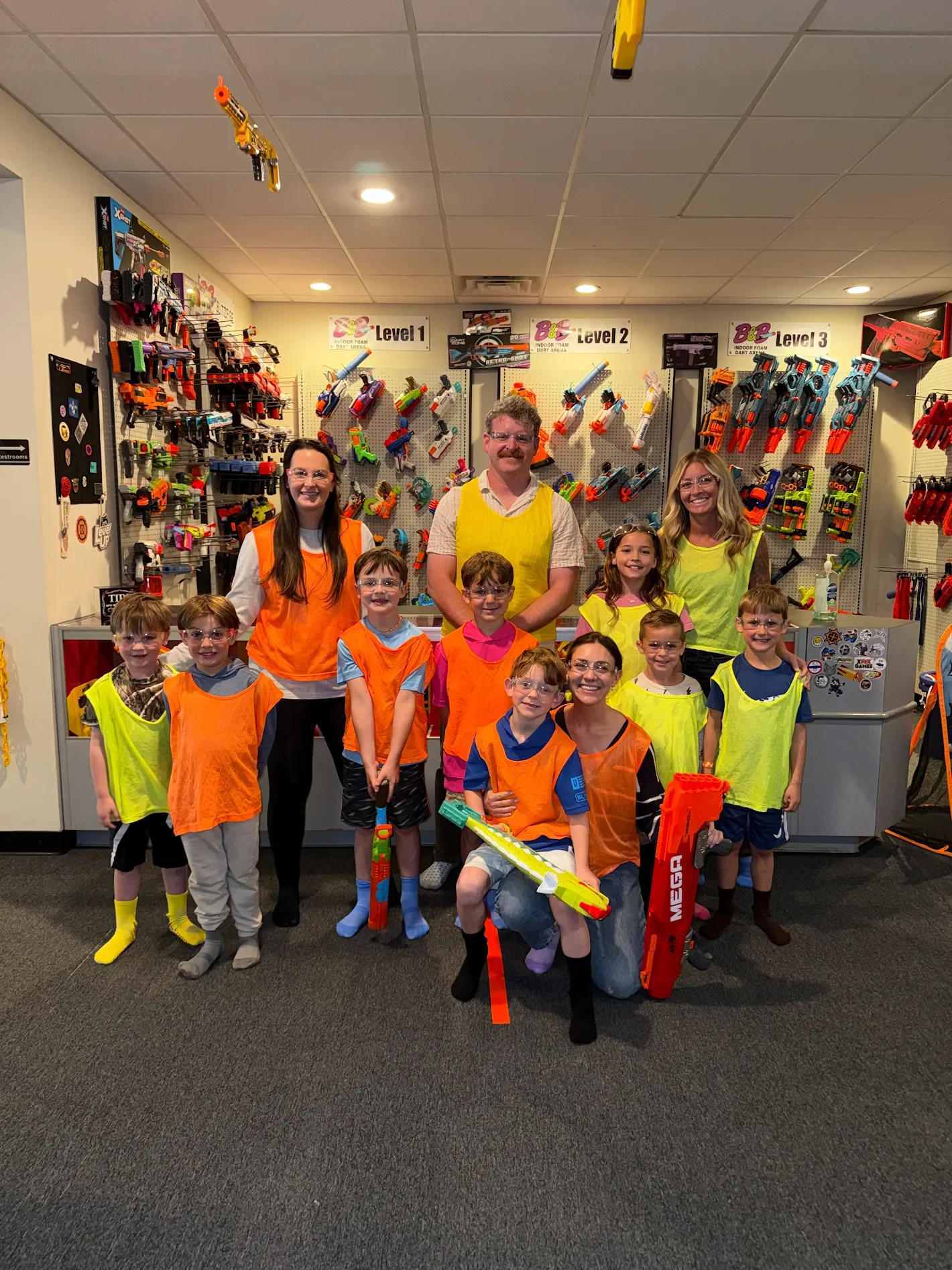 Group of children and adults posing with toy Nerf guns in a store, wearing colorful vests, with shelves of Nerf blasters labeled Level 1, Level 2, and Level 3 in the background.