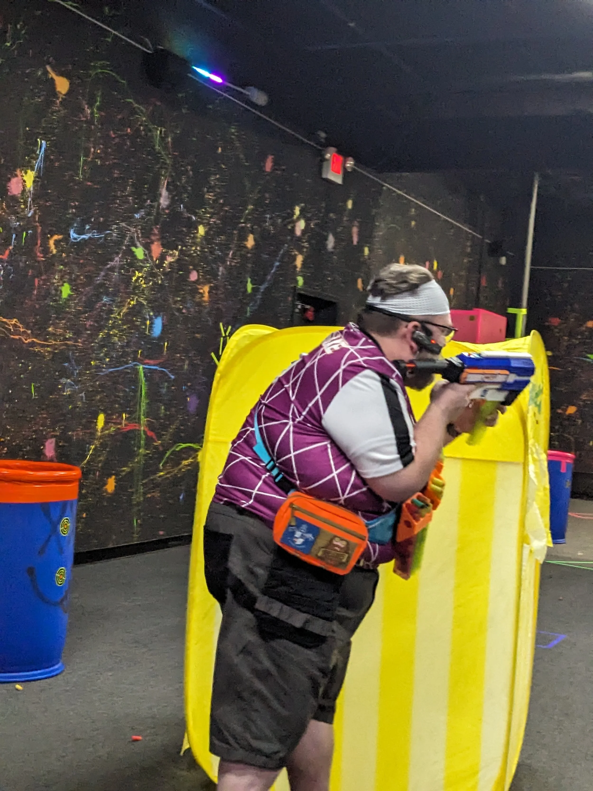 Person playing nerf, wearing a headband with sensors and aiming a nerf gun, with a colorful, abstract background and a yellow barrier