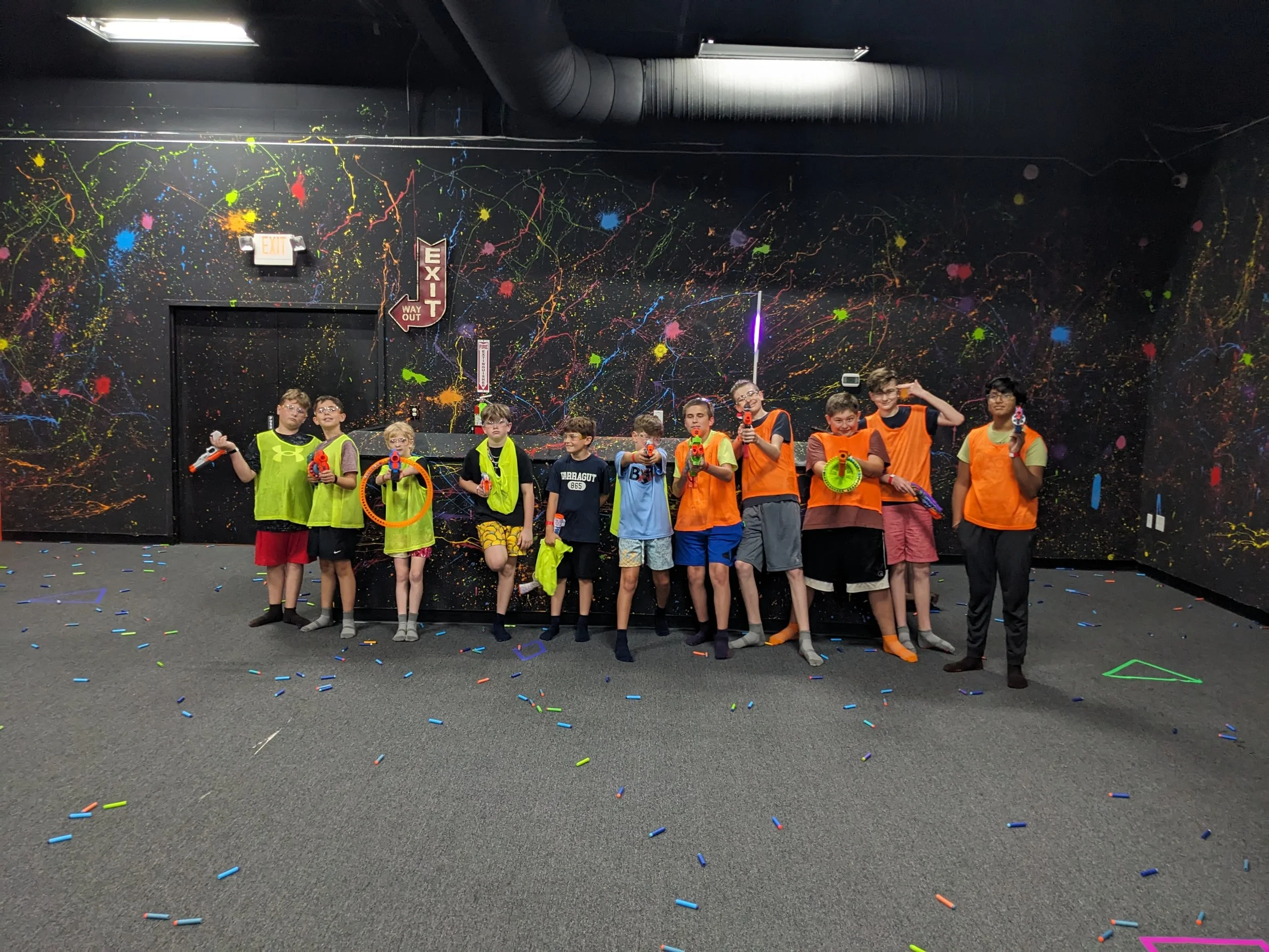 Group of kids in neon and orange vests holding water guns, standing in front of a black wall with colorful splatter paint and space-themed decorations, with confetti on the floor.