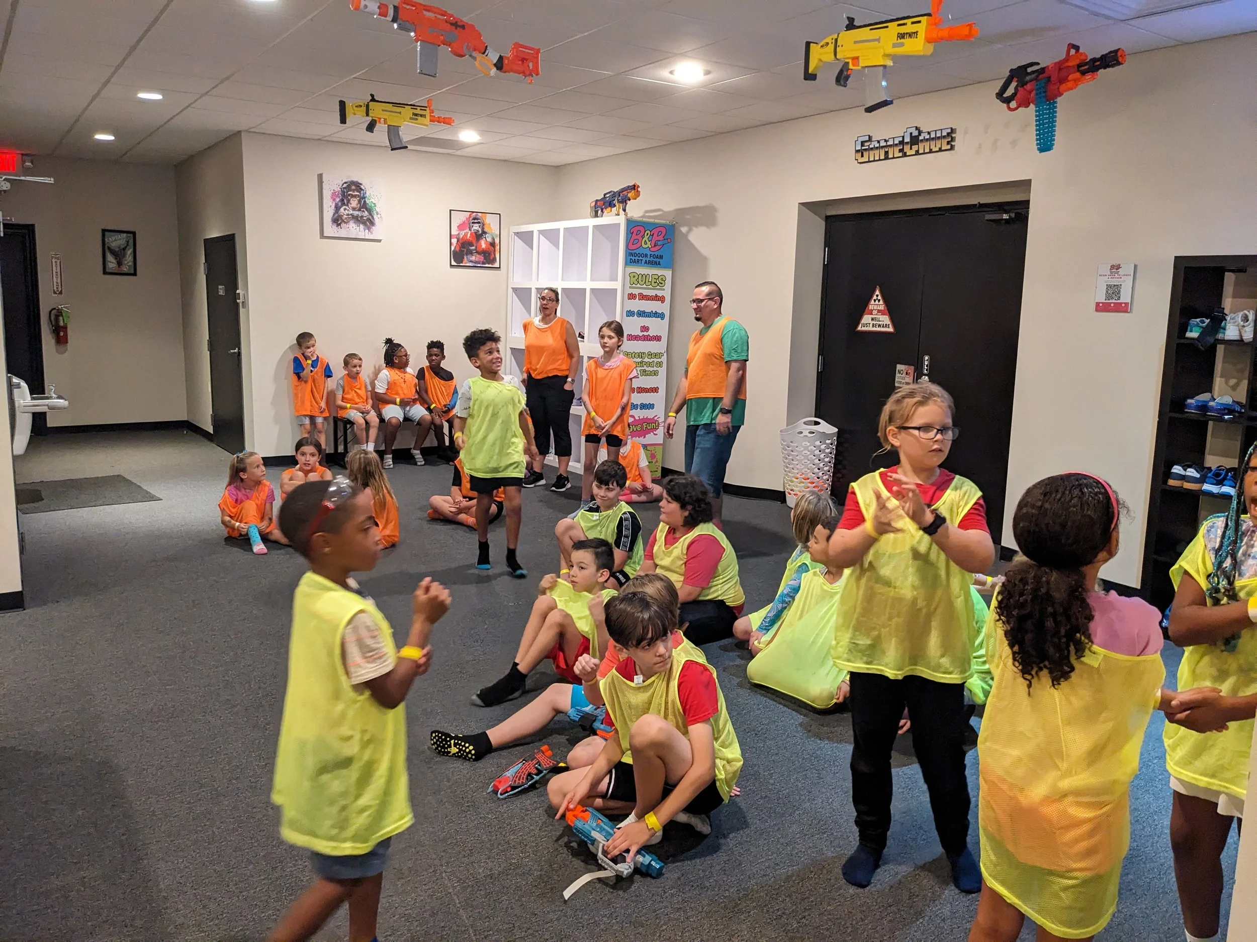 Children playing laser tag in an indoor arena with foam dart guns hanging from the ceiling, some children sitting on the floor while others stand or walk around, all wearing bright vests, with adults supervising in the background.