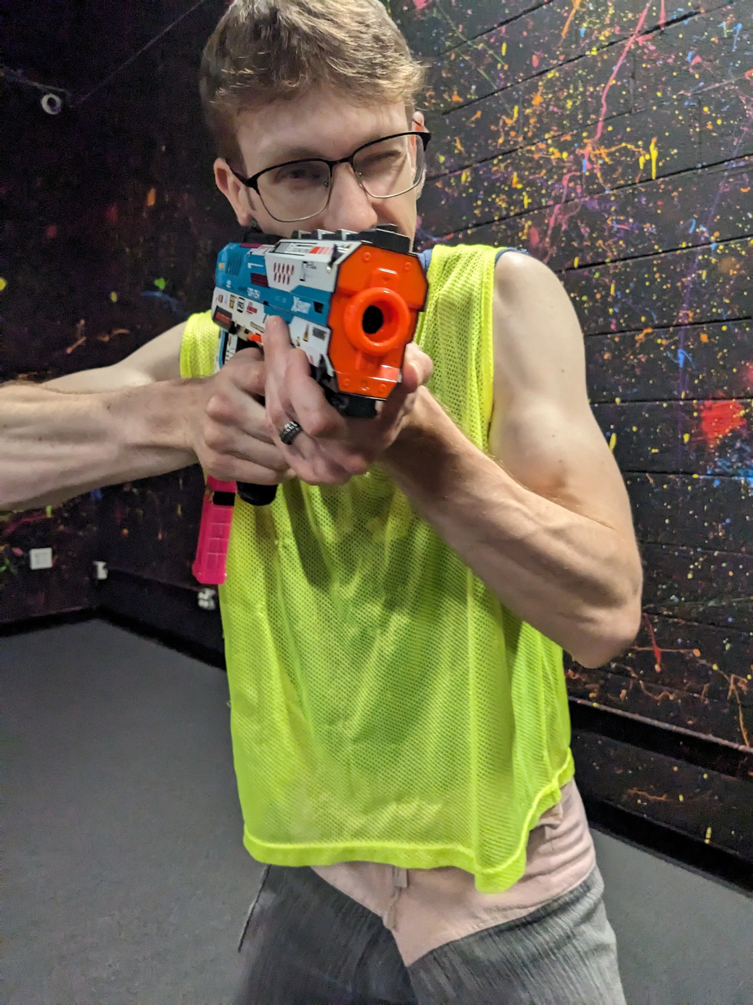 A person aiming a colorful nerf gun in an indoor nerf arena with a black wall splattered with colorful paint-like spots in the background.