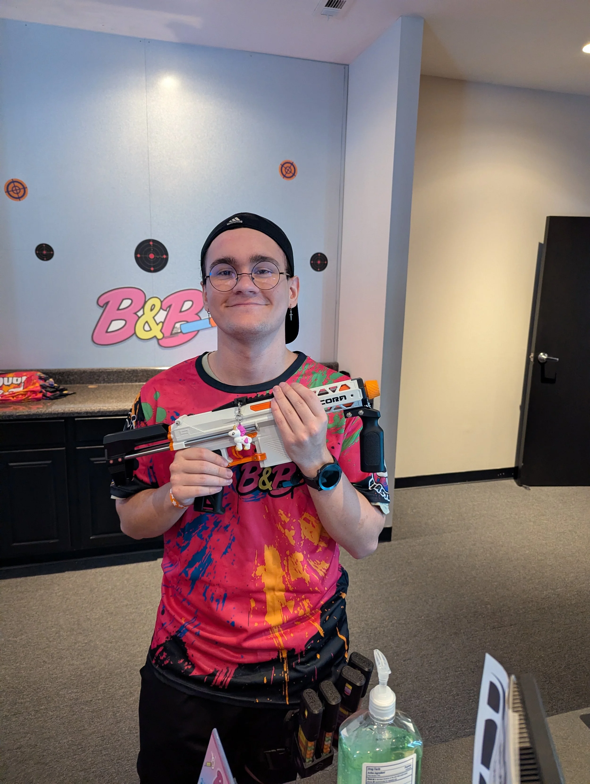 A young man wearing glasses, a black Adidas cap, and a colorful shirt holding a Nerf gun. There are target posters on the wall behind him, and a table with hand sanitizer, and various objects.