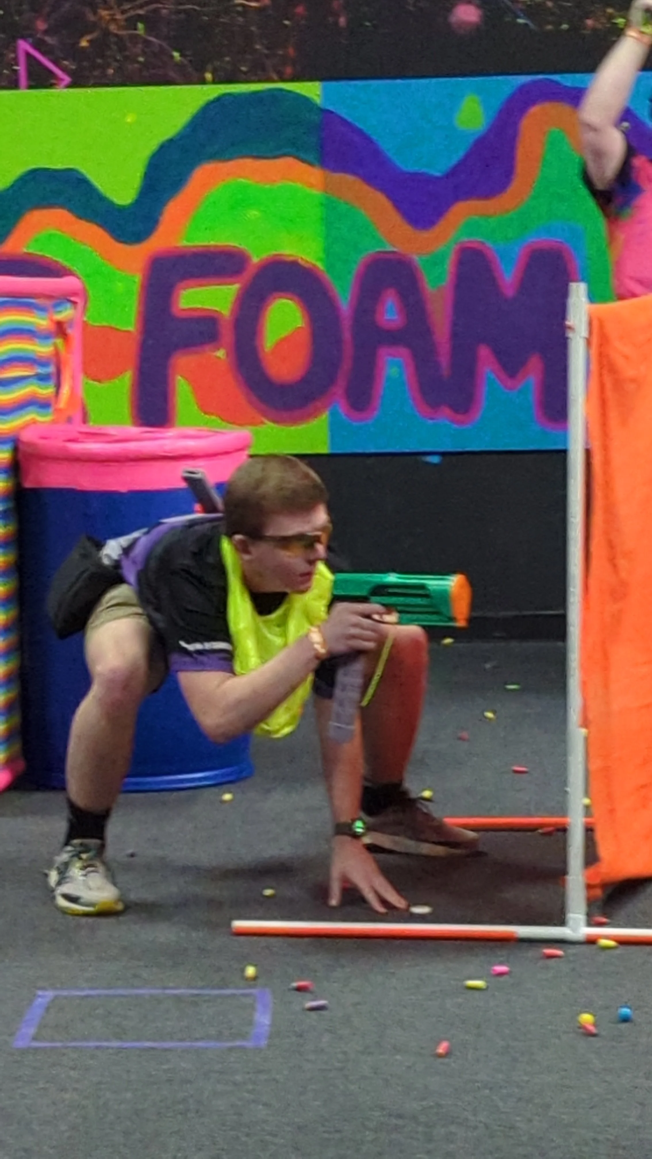 A kid in a crouched position holding a toy gun, playing a game of paintball or nerf warfare at an indoor event with colorful decorations, including a large vibrant sign that reads 'FOAM' in the background.