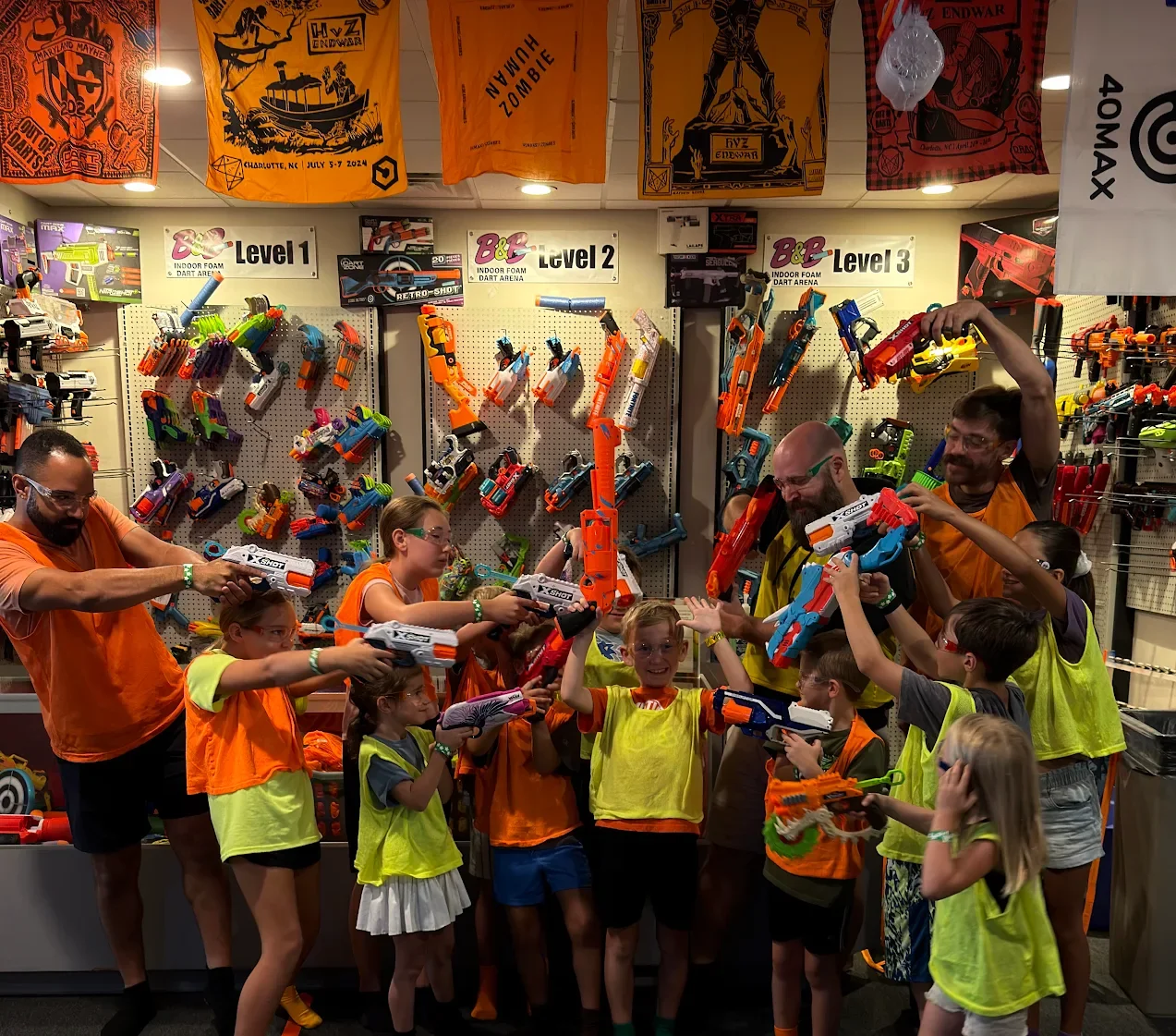 Children and adults in a nerf arena playing with large nerf guns, aiming at each other, surrounded by shelves filled with colorful nerf guns and darts, with signs indicating different levels of the dart arena.