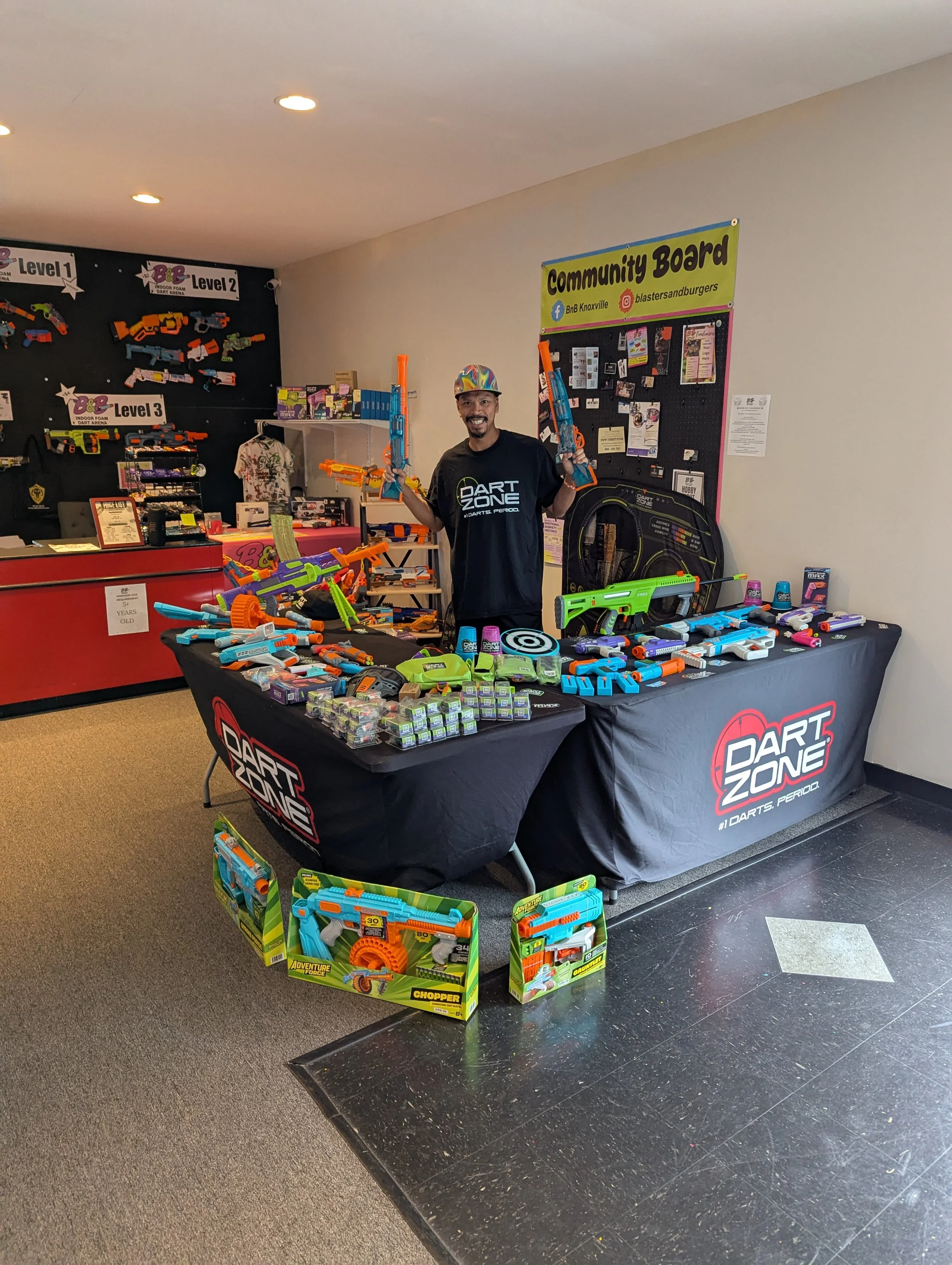 A man wearing a black T-shirt and multicolored cap standing behind a table filled with brightly colored toy dart guns at a Dart Zone store. There is a black board in the background with various dart guns displayed and a green sign that reads "Communi