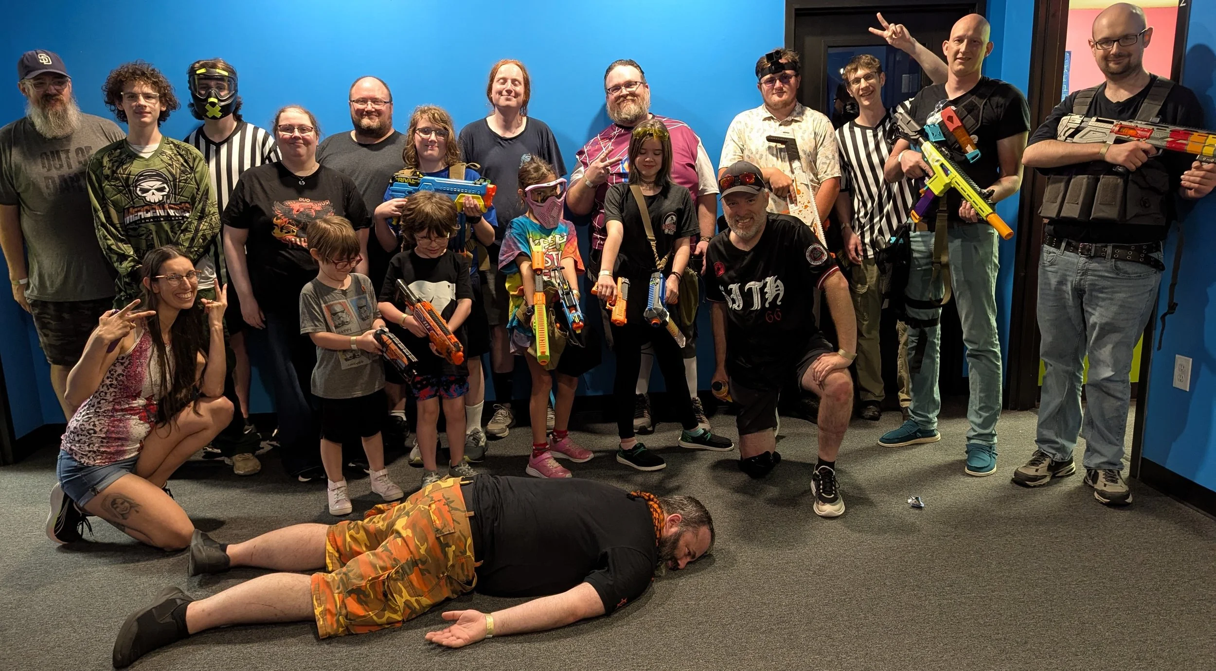 Group of people, including children and adults, posing with Nerf guns indoors against a blue wall. Some are holding nerf guns, others make peace signs or pose. One person is lying on the floor.