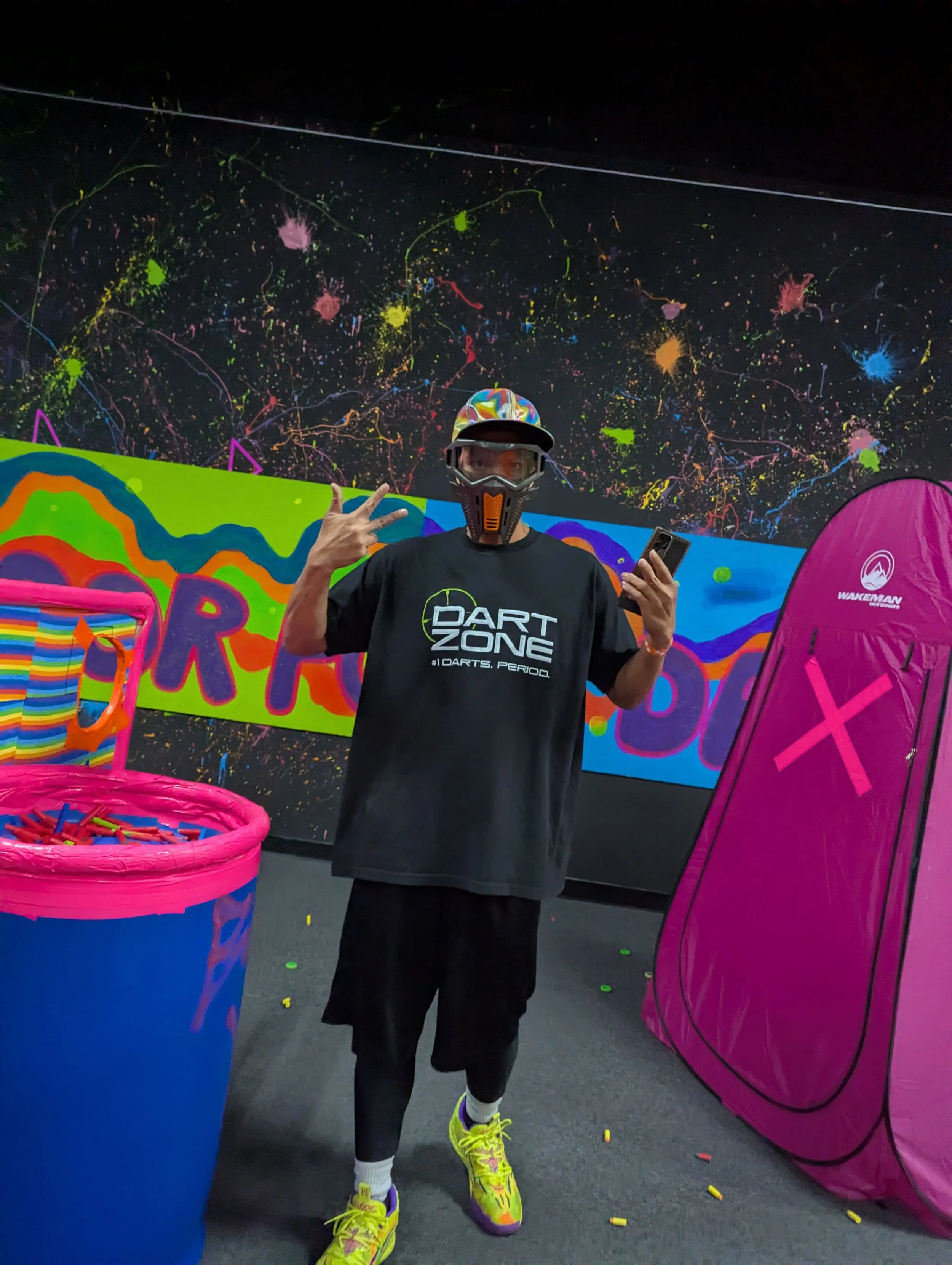 A person wearing bright yellow and purple athletic shoes, black athletic shorts, and a black T-shirt with words and graphics stands in a bowling alley. The individual is wearing protective eyewear and a colorful helmet, posing with a peace sign while