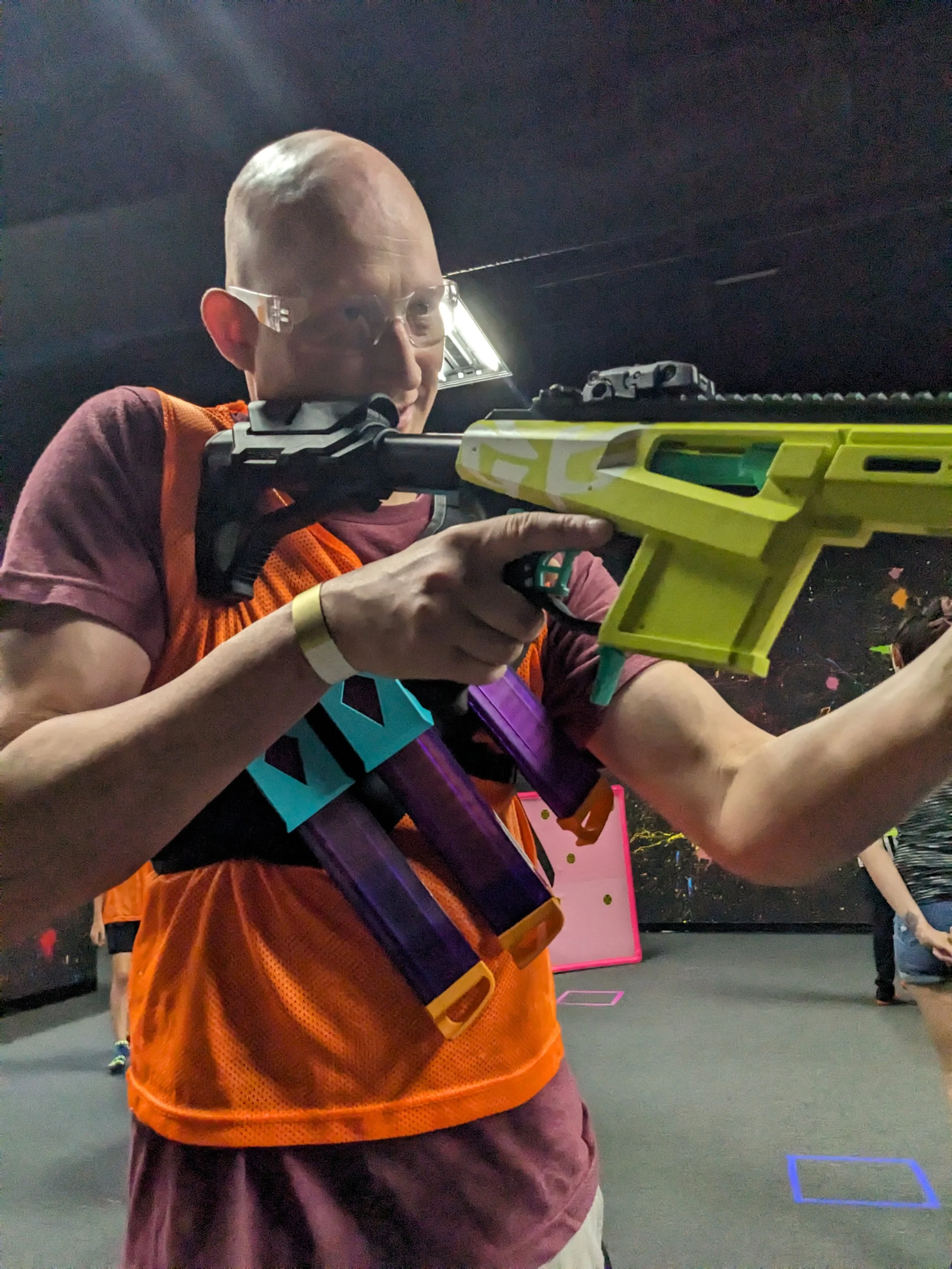 Person wearing safety glasses aiming a yellow and black nerf gun in an indoor arena.
