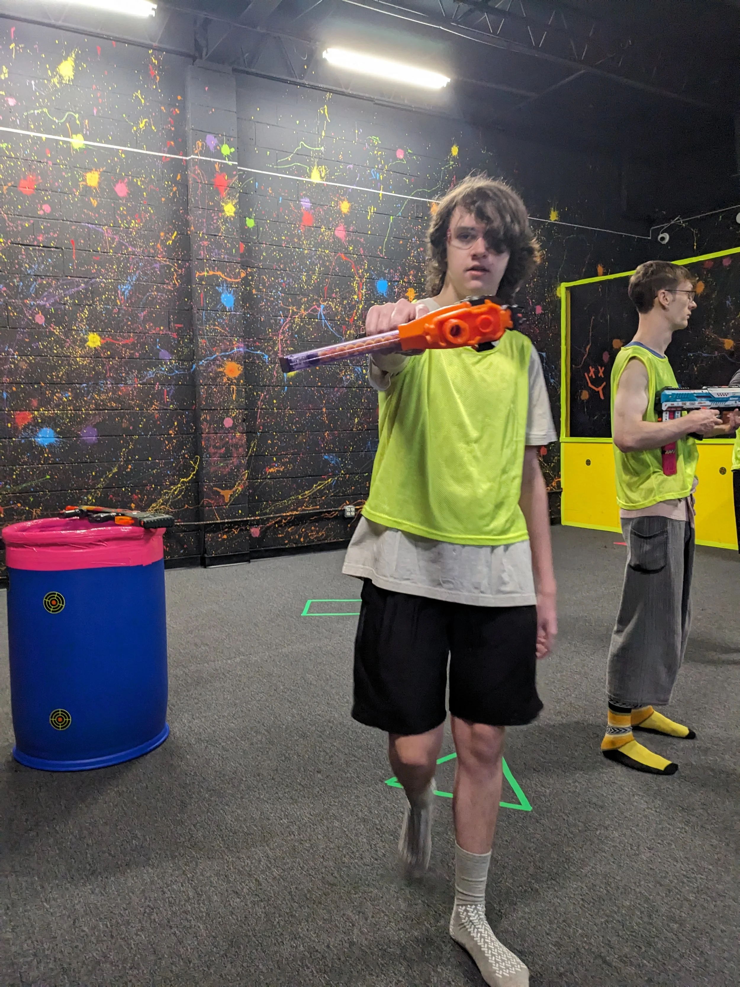 Young man with curly hair holding a nerf gun in an indoor arena with a colorful splatter paint wall.