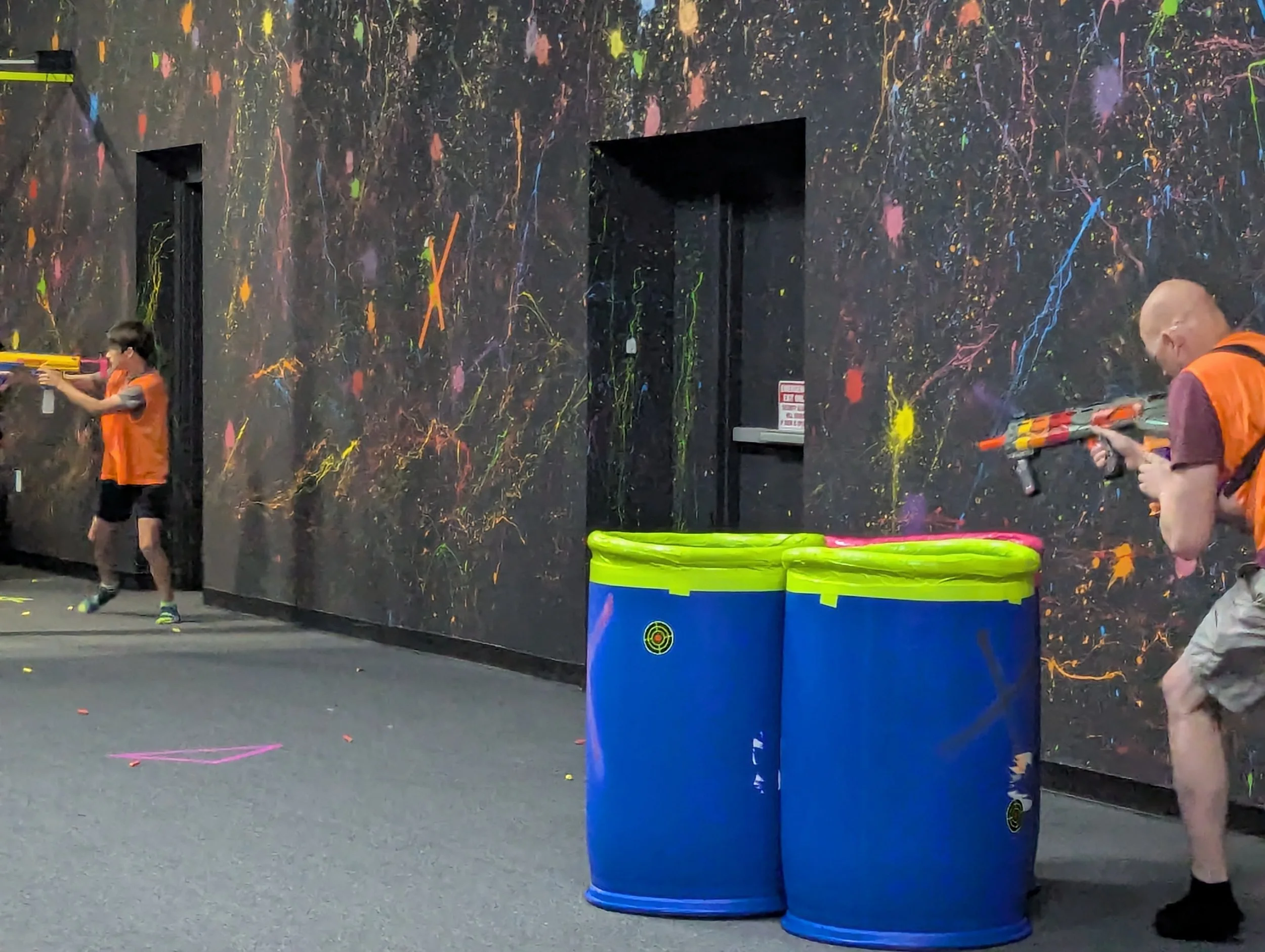 Two kids and an adult playing laser tag with toy guns in an indoor arena with colorful splatter paint walls, and three blue barrels in the foreground.