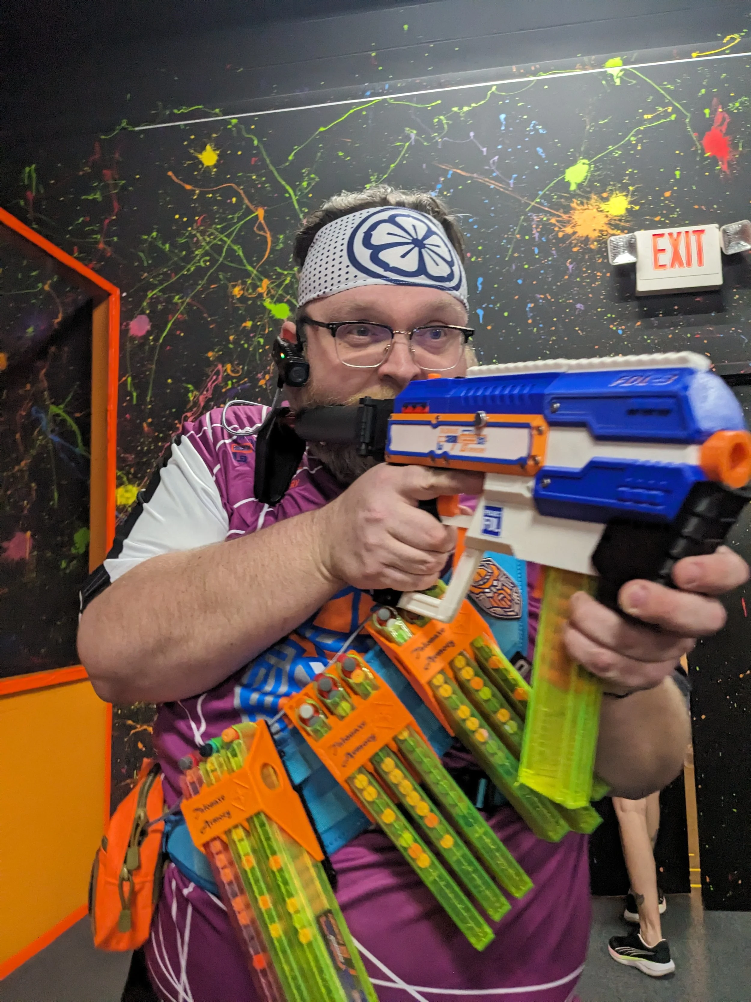 A man dressed in colorful Nerf-themed attire aiming a Nerf blaster at a laser tag or Nerf arena with a black wall decorated with splashes of bright colors and an orange and white exit sign in the background.
