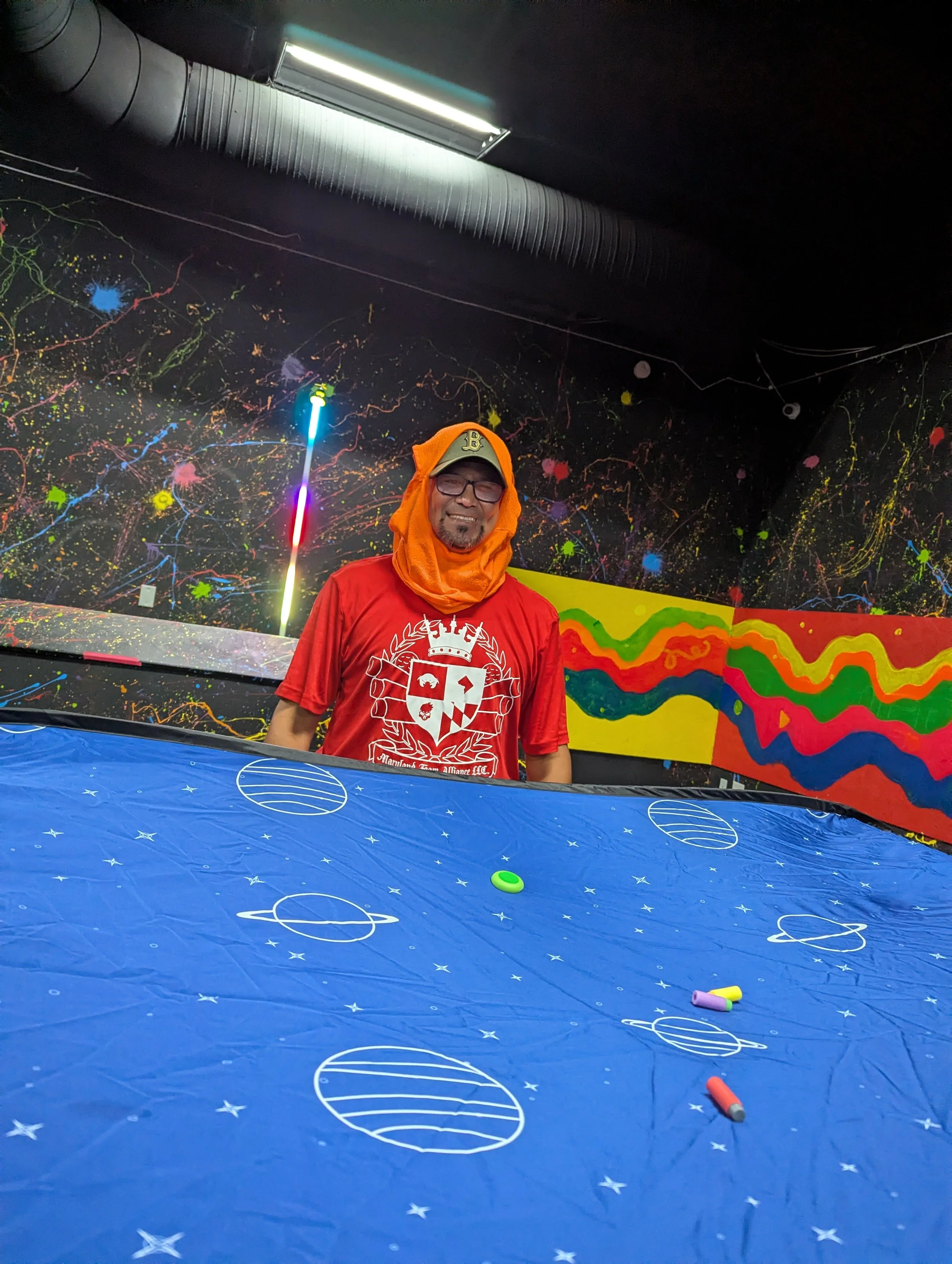 A man smiling, wearing glasses, an orange hoodie with the hood over his head, a beige baseball cap, and a red shirt, standing in a colorful, space-themed room with black walls decorated with bright paint splatters and abstract designs, and a blue tab