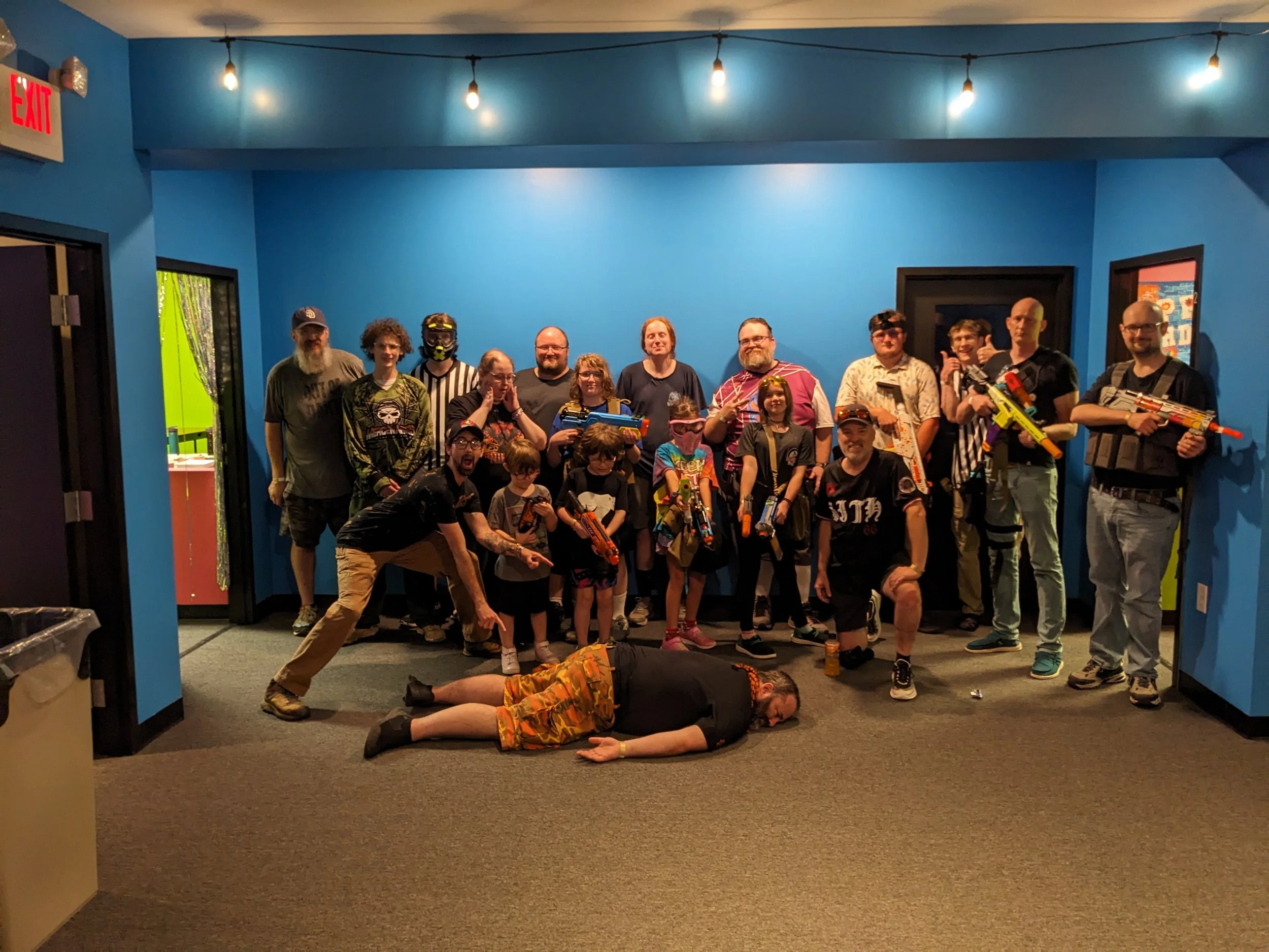 Group of people, including children and adults, posing indoors with toy guns, some wearing costumes and accessories, one person lying on the floor, in a blue-walled room with lighting string overhead.