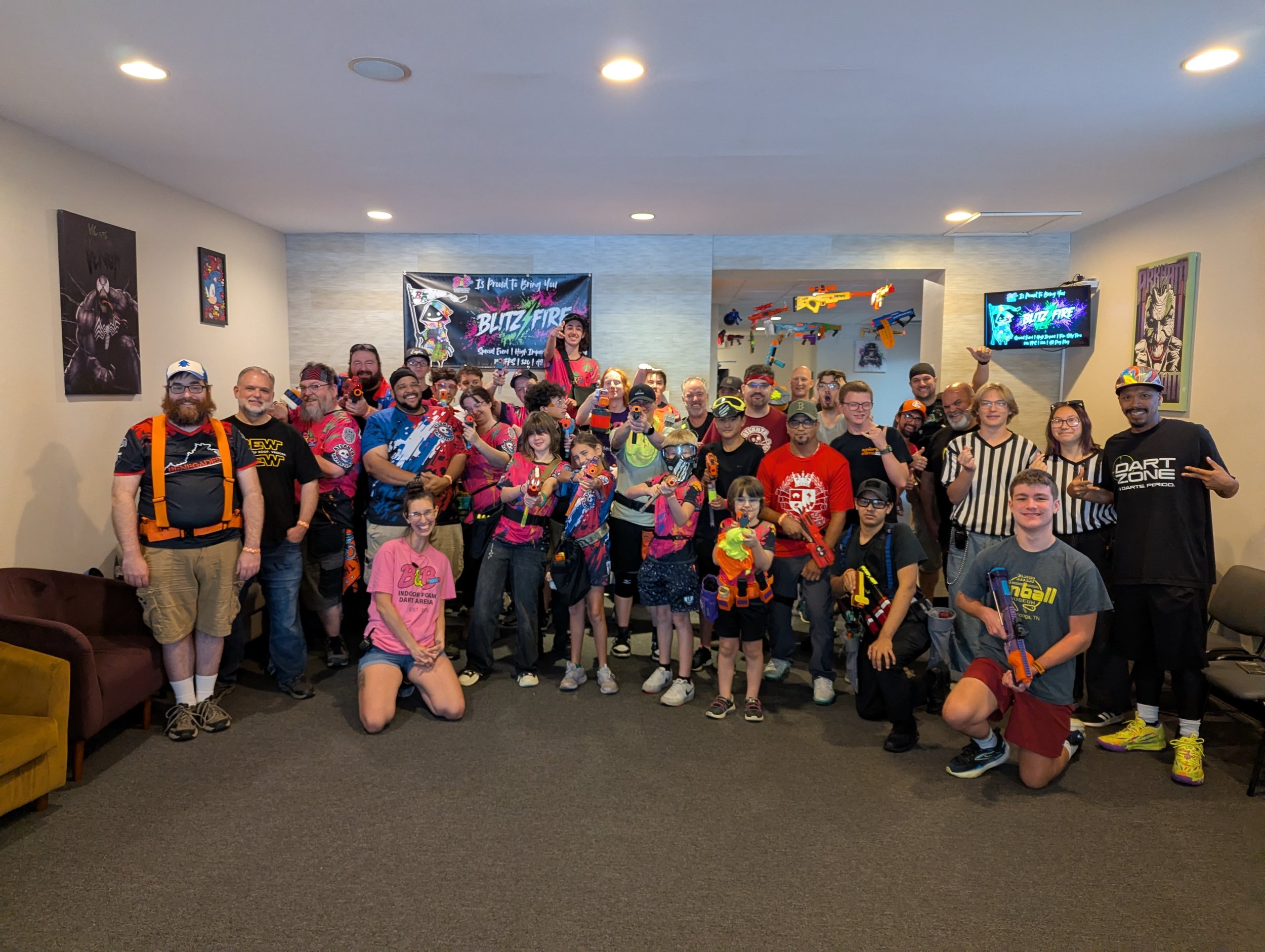 Group photo of people, mostly children and adults, holding toy guns, inside a recreational facility with posters and painted walls.