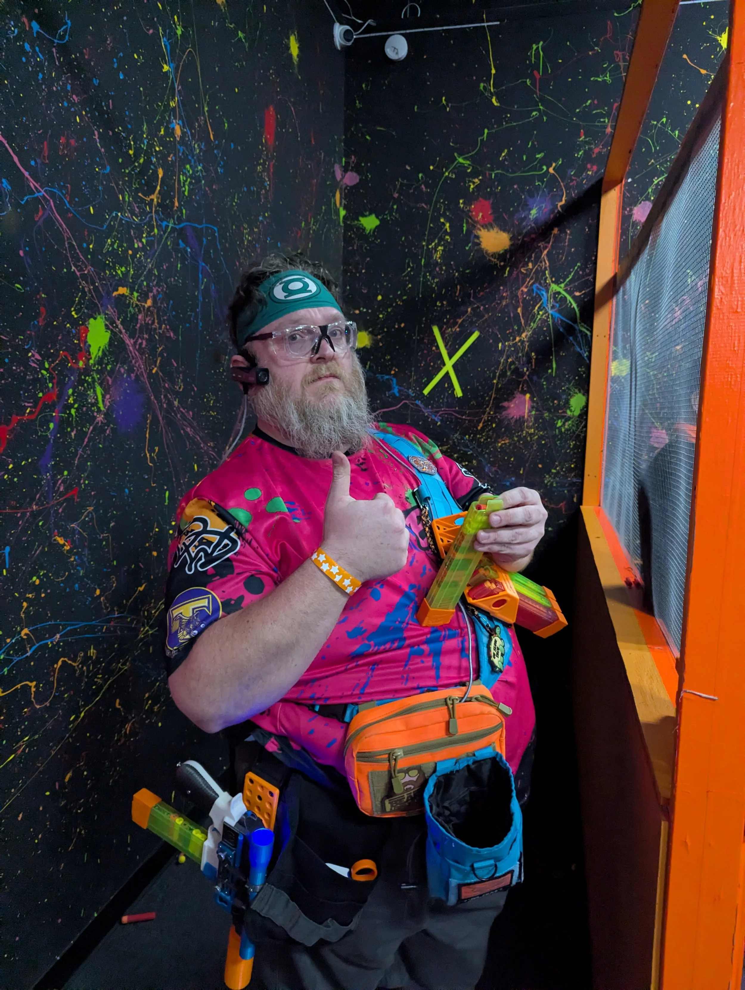 A man with a beard wearing glasses, a headband, and a colorful shirt, giving a thumbs-up while standing in a room with black walls splattered with colorful paint. He has a small orange and blue bag around his waist and is holding a toy blaster with n