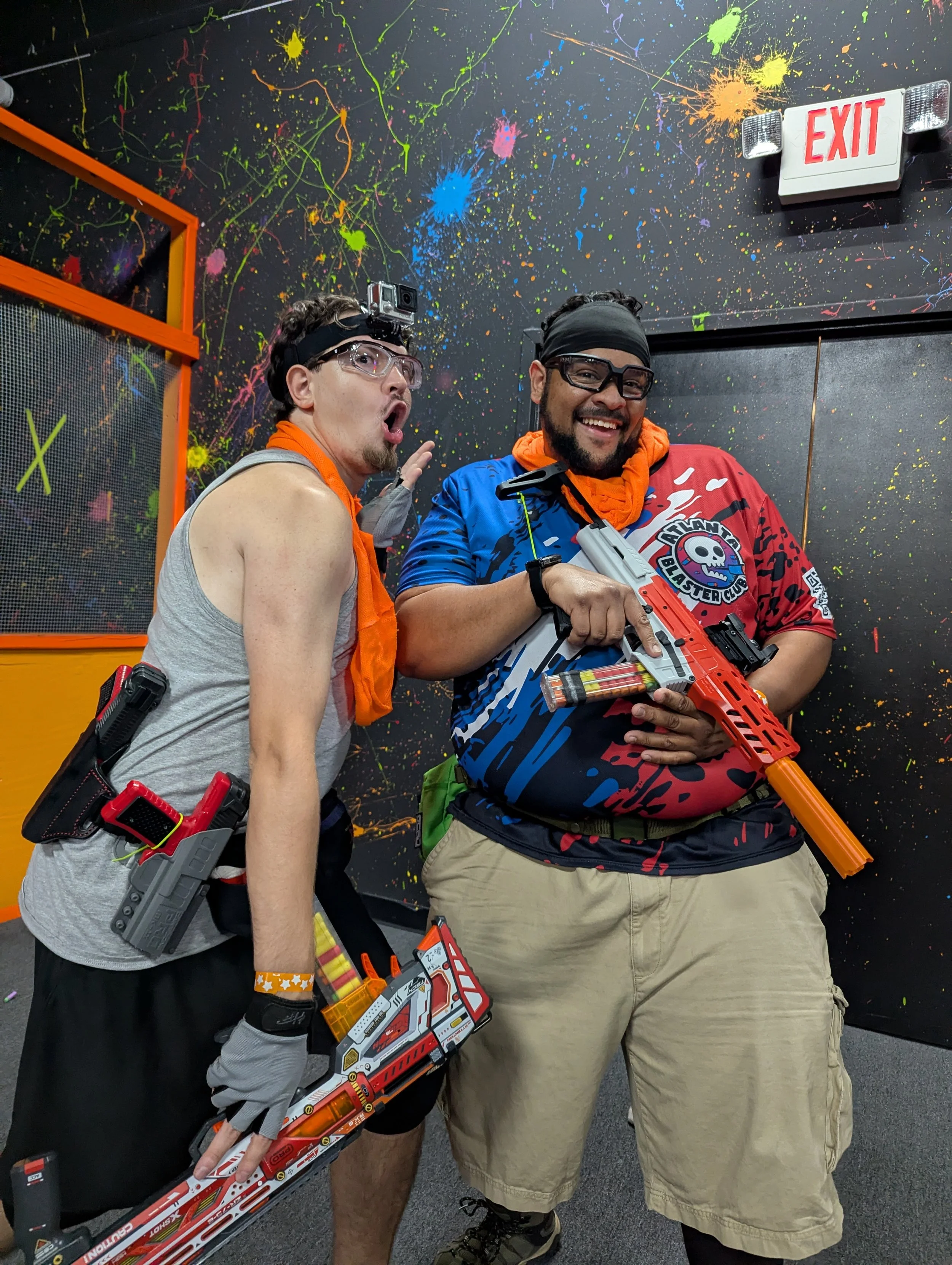 Two men posing with toy guns in front of a black wall with colorful splatter paint design. One man is wearing glasses, a headlamp, and a gray tank top. The other man is smiling, wearing glasses, a black headband, and a multicolored shirt with a skull