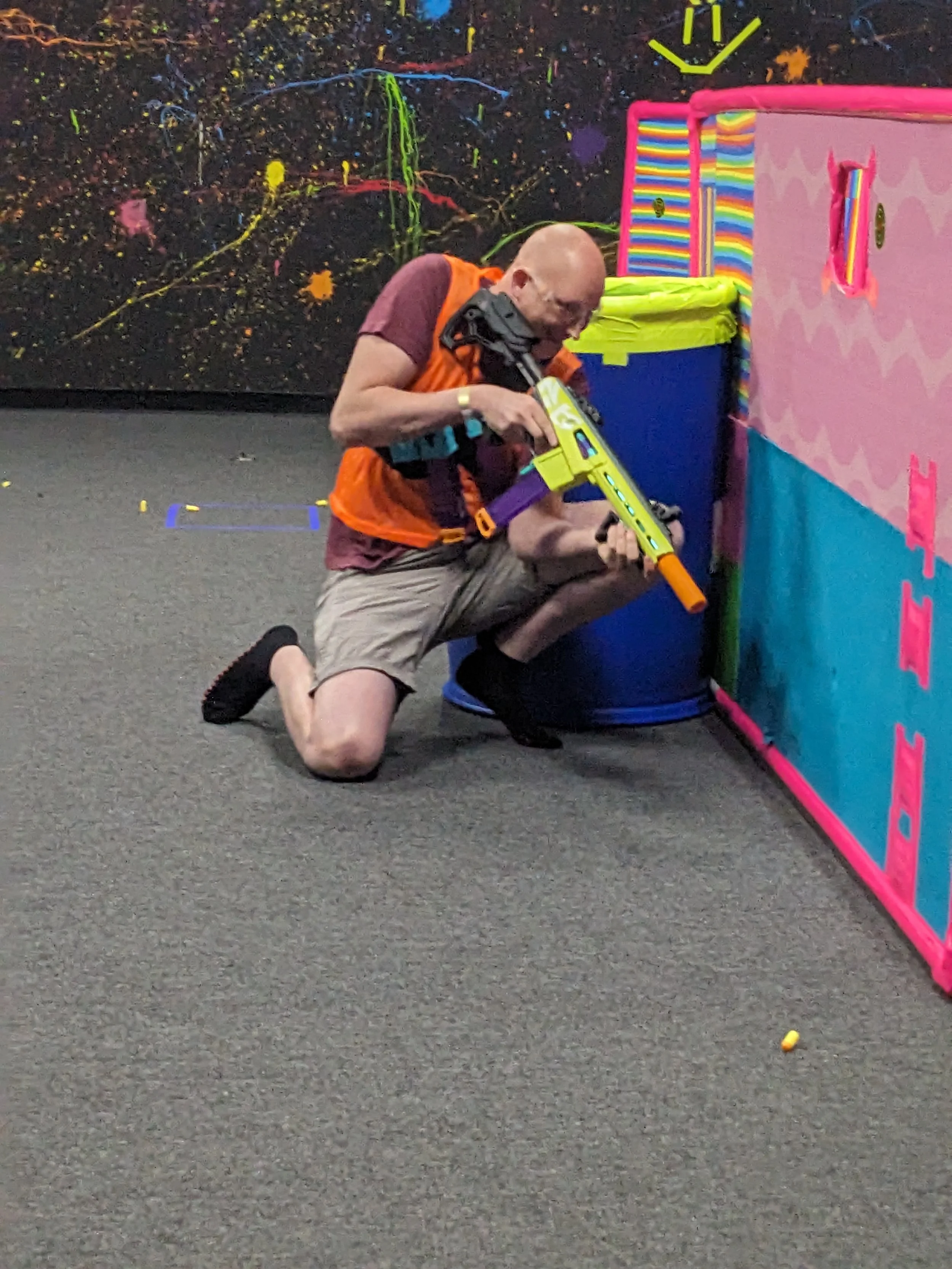 A man kneeling on one knee, holding a toy nerf gun, aiming it behind a pink and blue foam wall in an indoor nerf arena, with colorful decorations in the background.