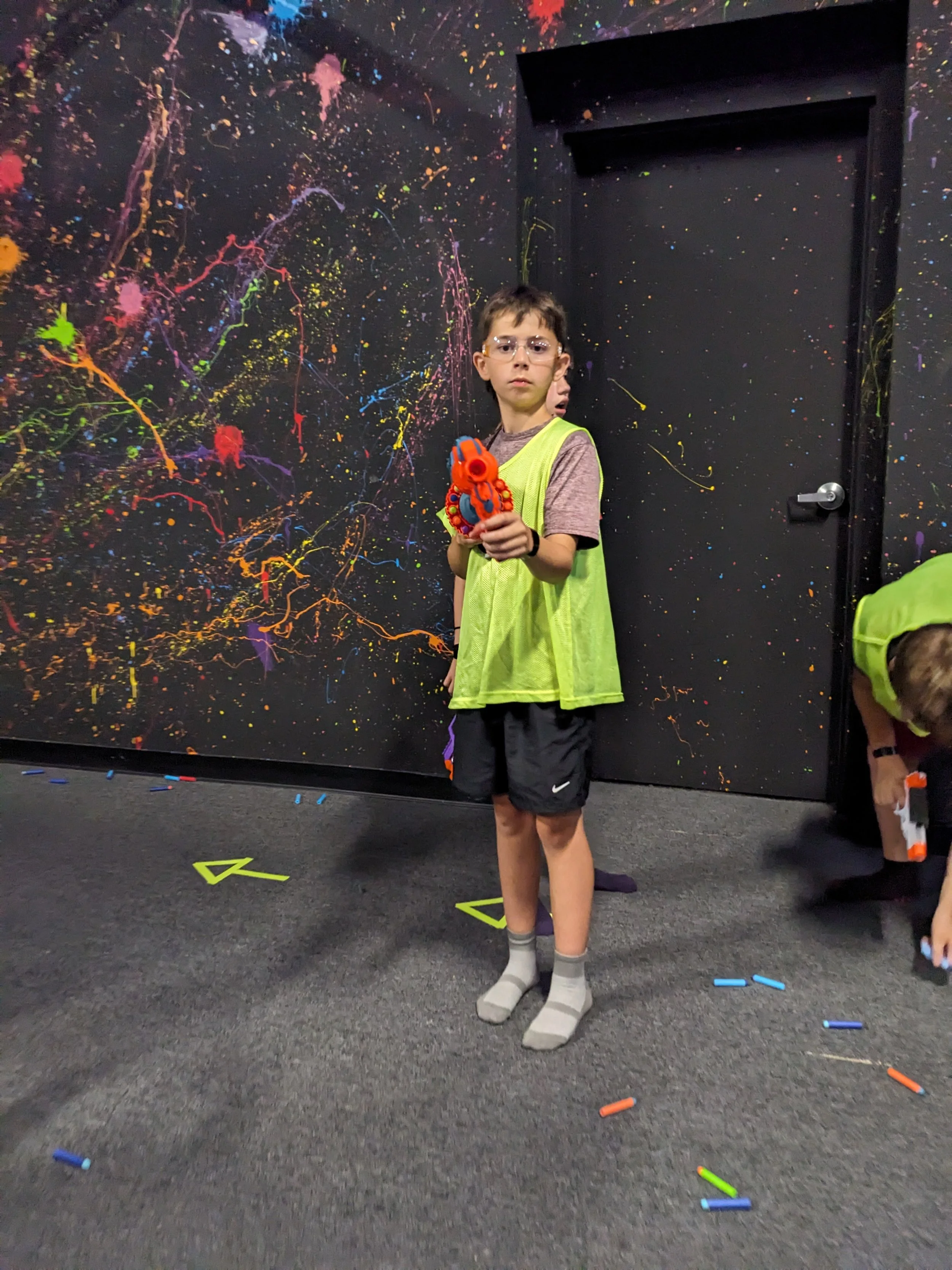 Boy in yellow vest holding a toy water gun, standing on a carpeted floor with scattered toy darts in front of a black wall with colorful paint splatter art.