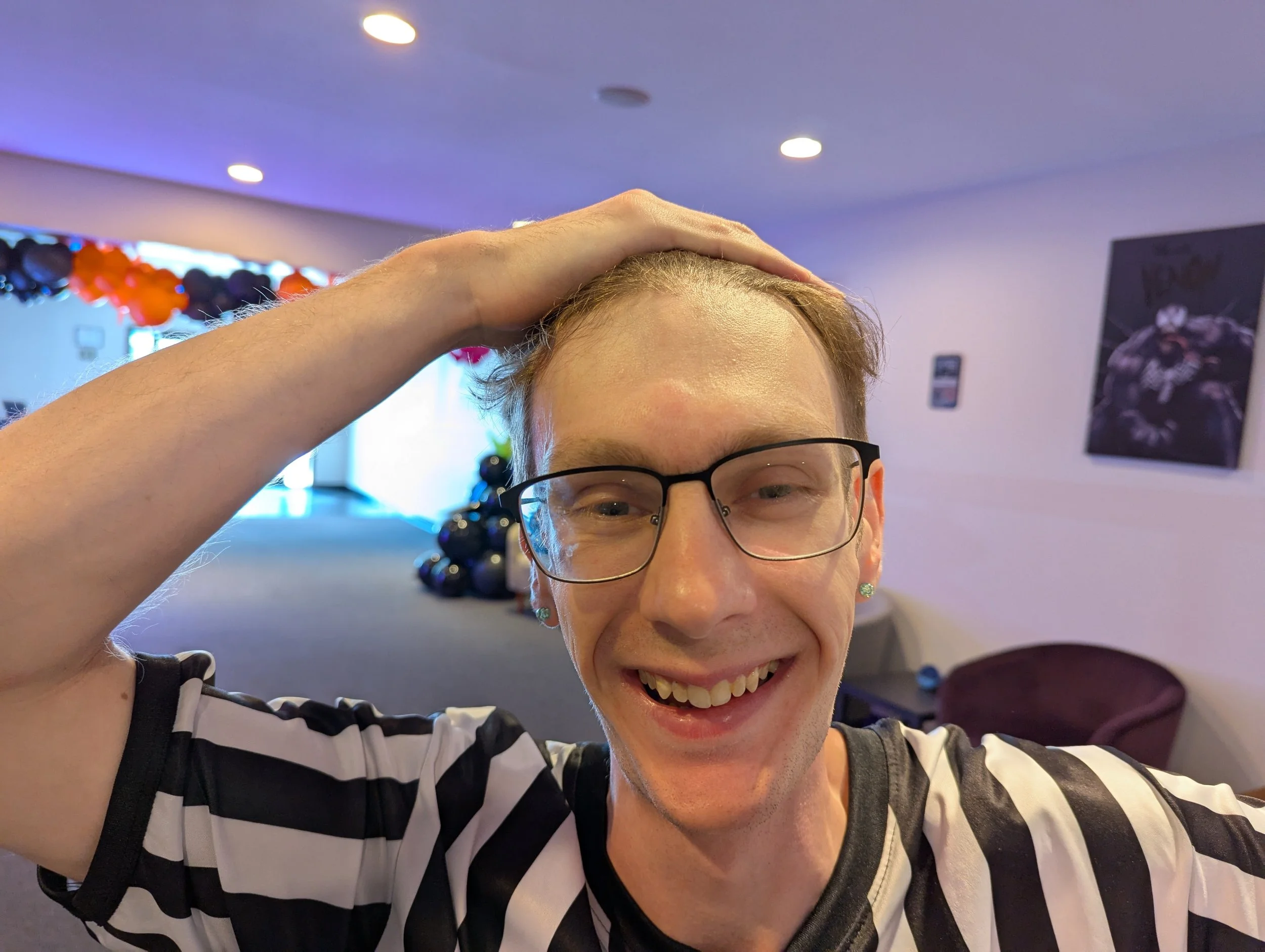 A smiling person with glasses, wearing a black and white striped shirt, holding their hand on top of their head inside a decorated indoor space.