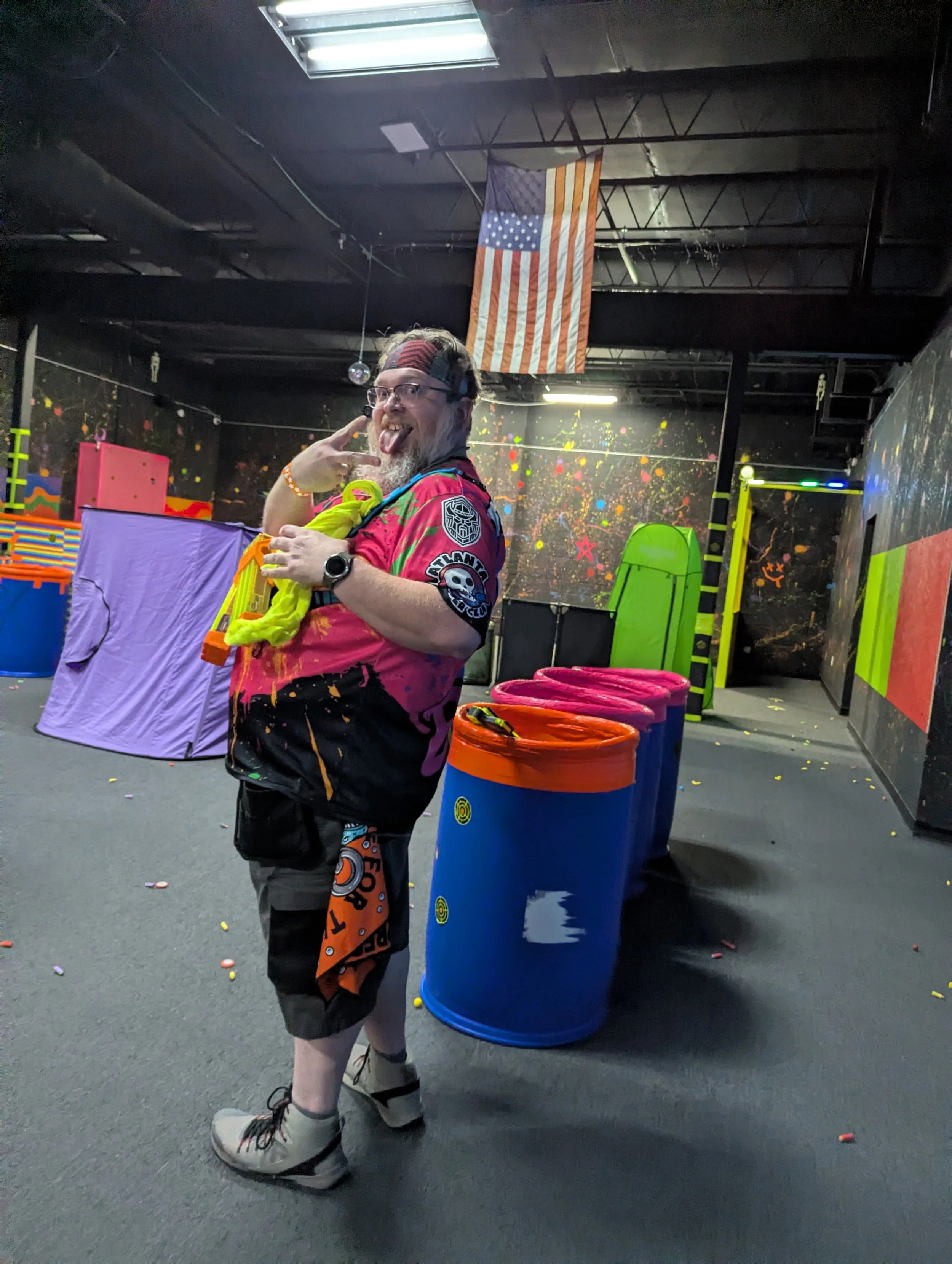 A man with glasses, a beard, and a flag headband standing in an indoor laser tag arena, making a playful face with his tongue out and hand gesture, surrounded by colorful foam barriers and neon-themed decorations.