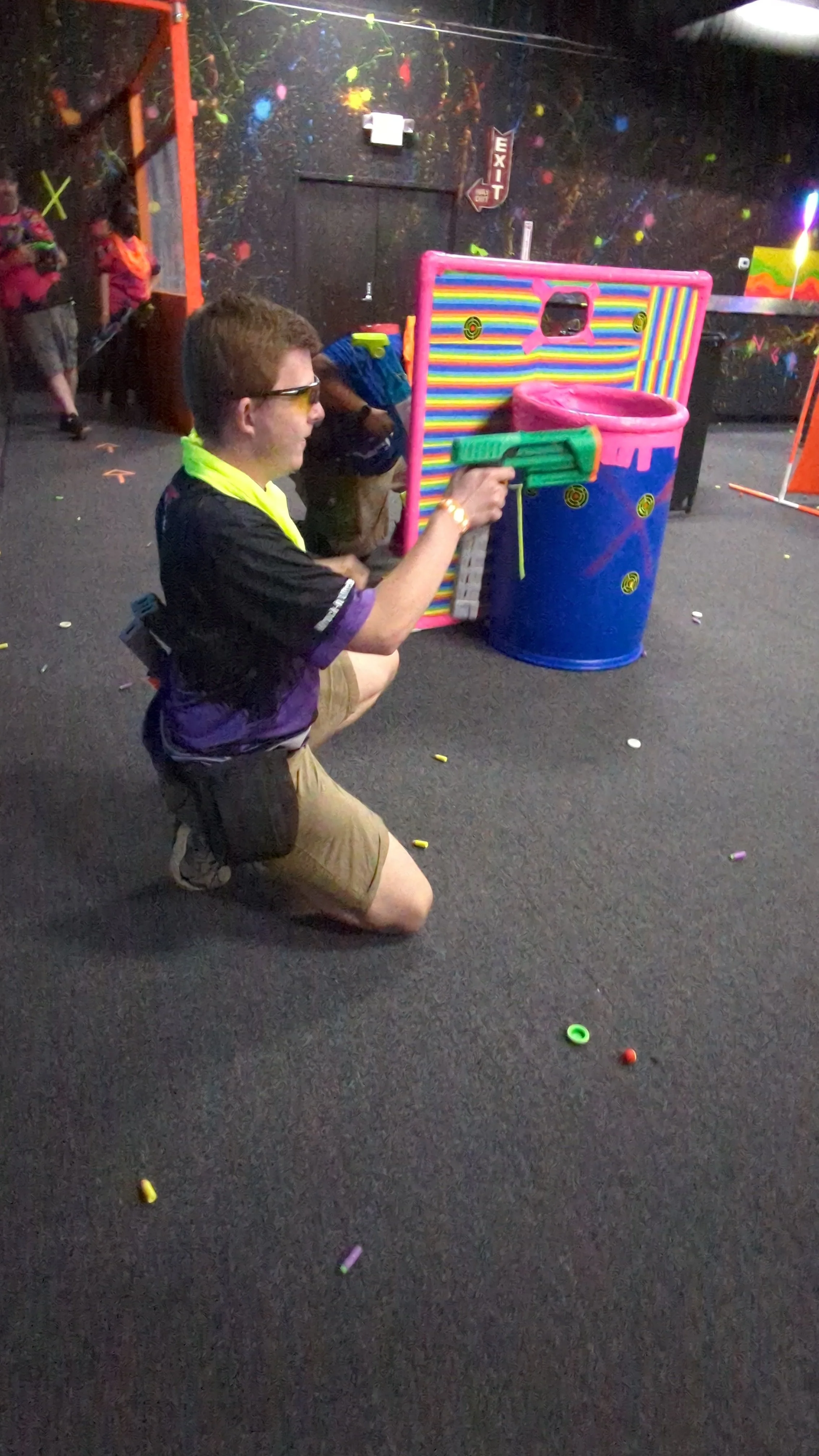 A man kneeling on the floor playing a game of indoor skee ball or a similar arcade game, aiming with a toy gun at a pink plastic basketball hoop and target station. The floor is scattered with small, colorful plastic tokens. The background includes a