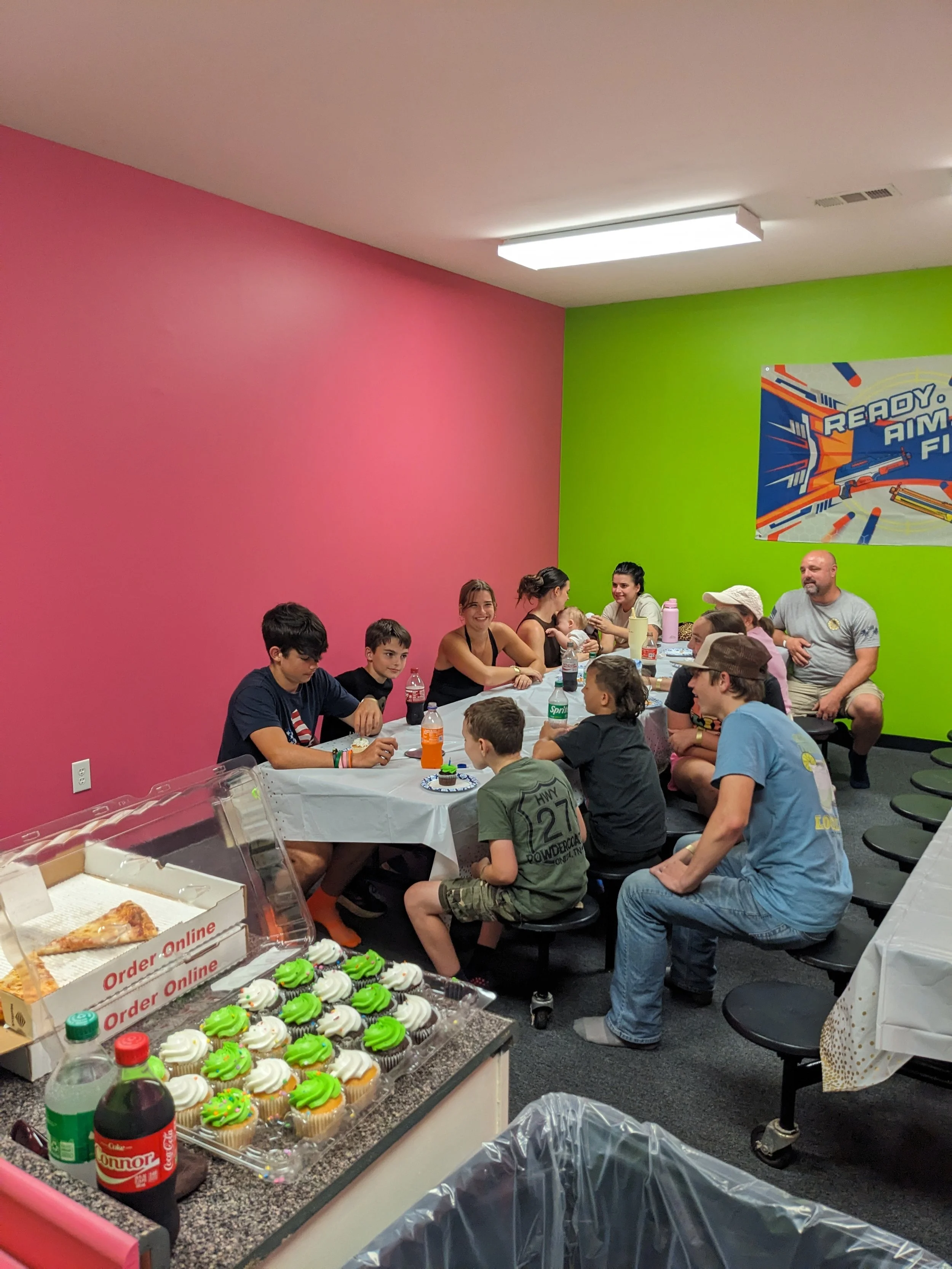 Children and adults sitting at a long table with cupcakes, drinks, and pizza in a brightly colored room celebrating a birthday, with pink and green walls and a party banner.