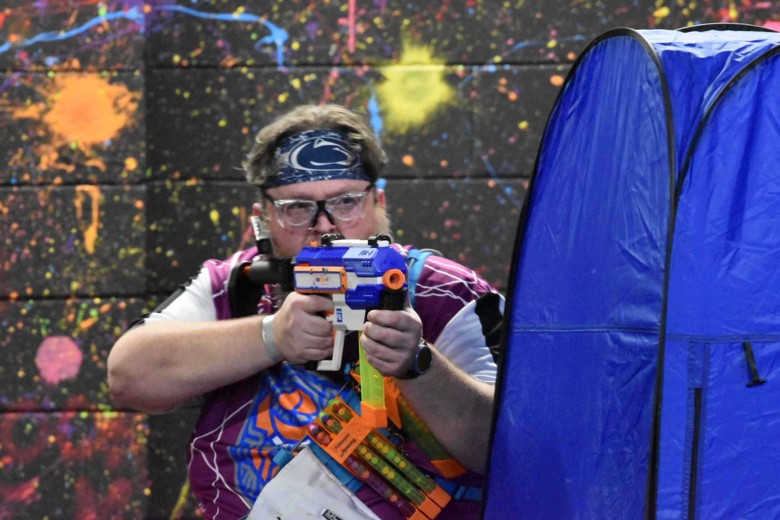 A man playing nerf aiming a nerf gun behind a blue cover, wearing a headband and glasses, with a colorful, splatter-painted background.