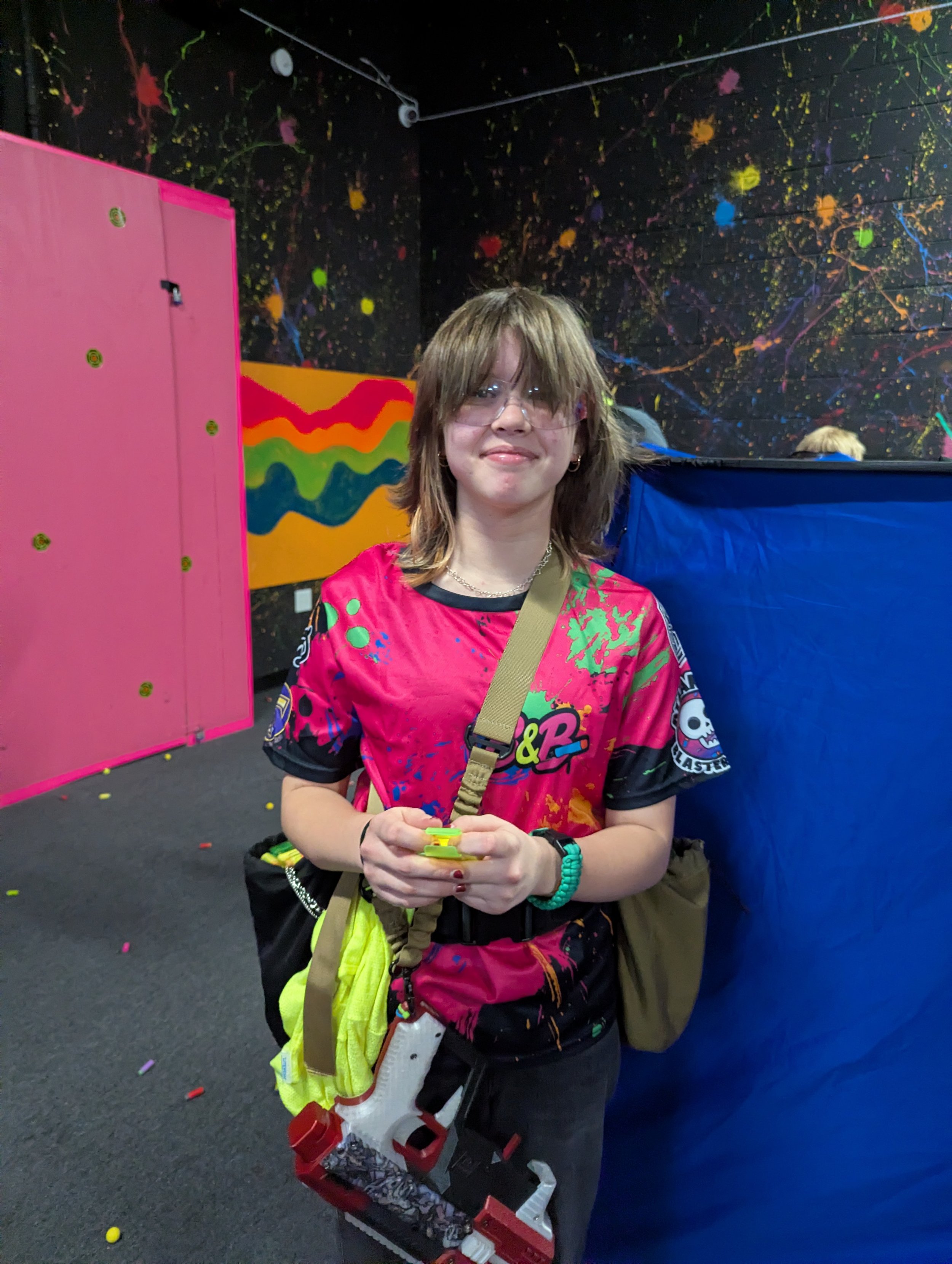 A person with shoulder-length light brown hair, glasses, and hoop earrings smiling, holding a toy cannon in front of a colorful abstract black wall with splashes of neon colors. They are wearing a vibrant pink, green, and black patterned T-shirt with