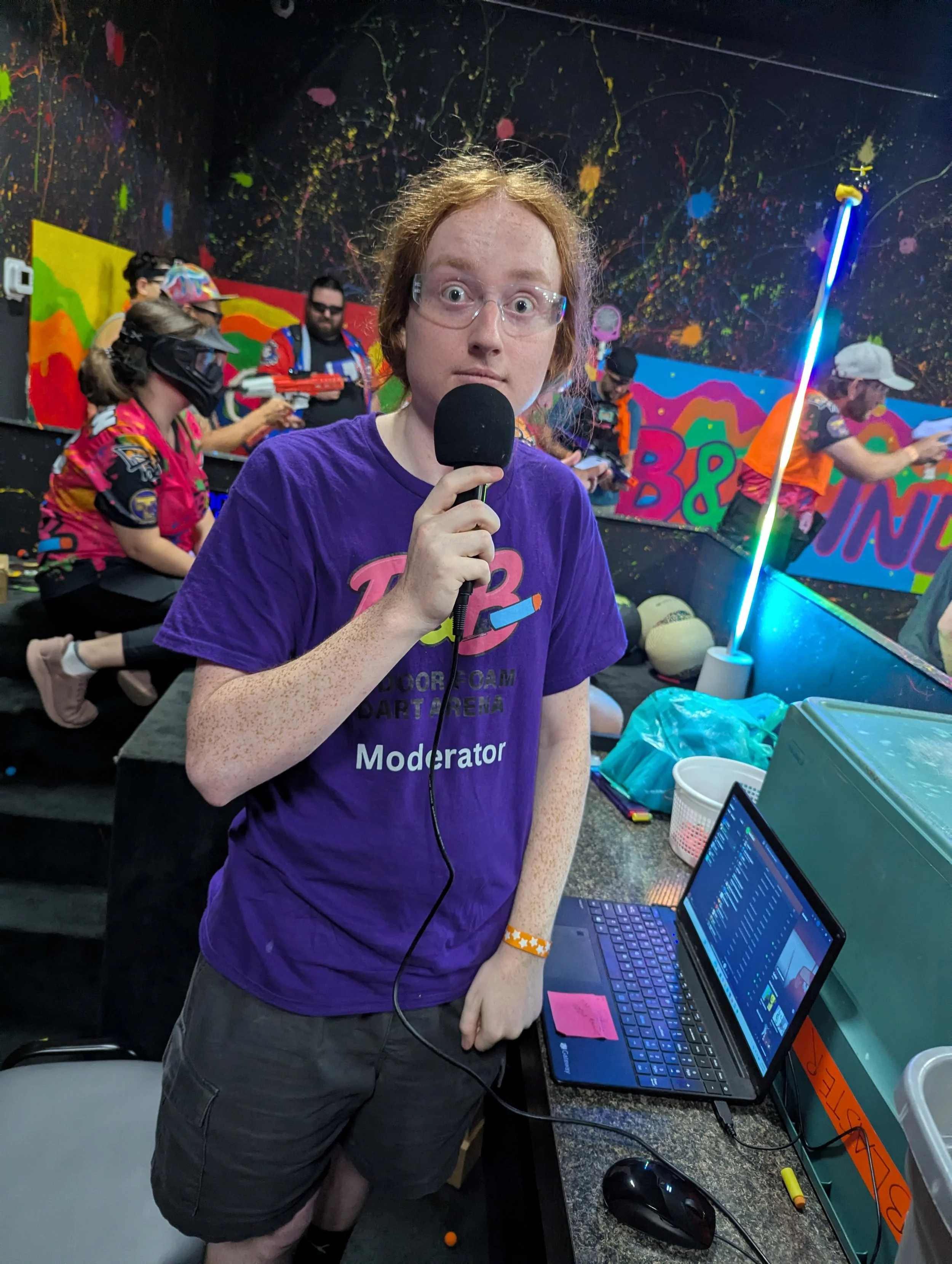 A person with red hair, freckles, and glasses holding a microphone and looking at the camera, standing in front of a colorful, splattered paint wall. The person is wearing a purple shirt that says "Moderator" and appears to be at an event with multip