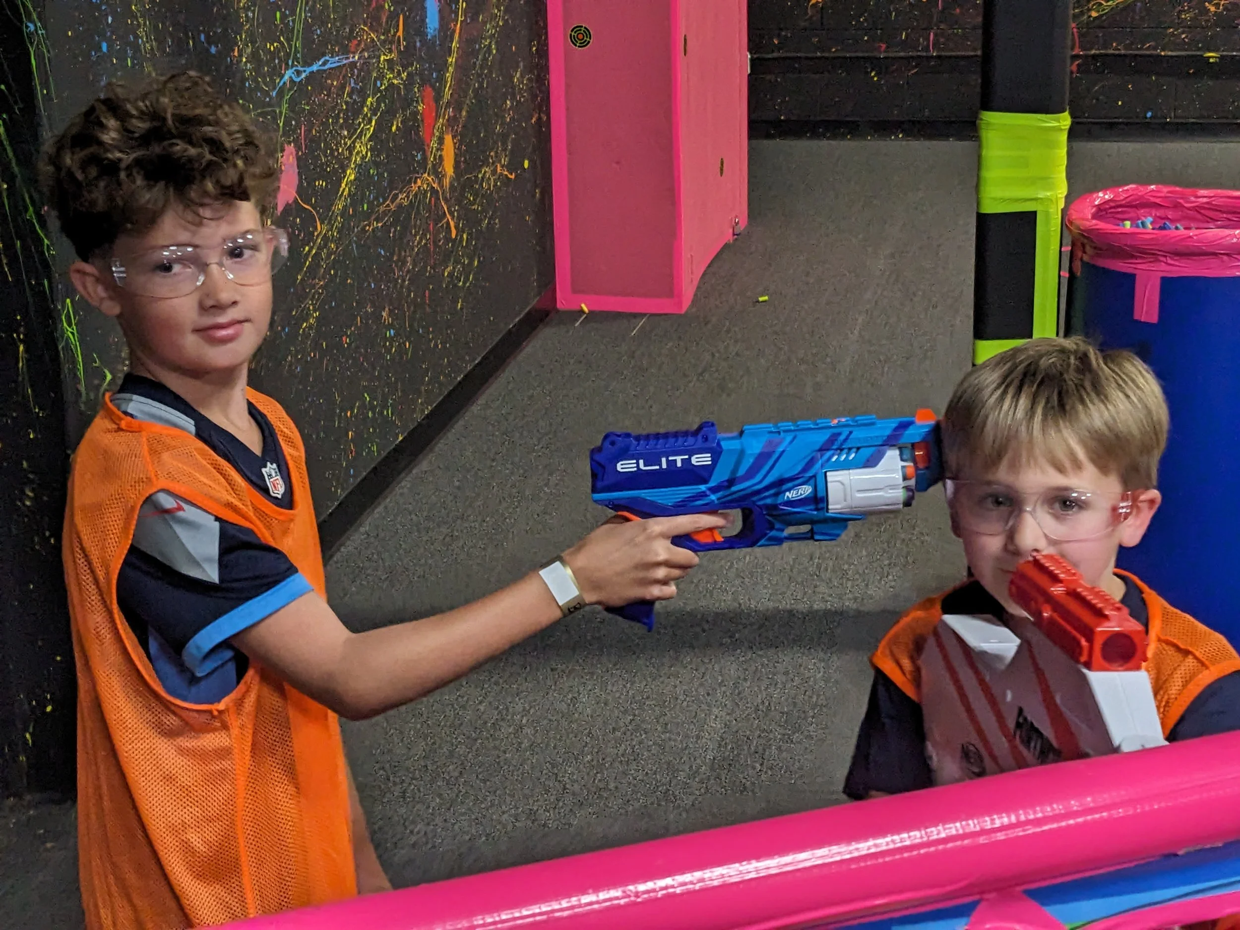Two boys wearing safety glasses and orange vests playing with toy guns in an indoor laser tag arena. One boy is aiming a blue gun at the other boy, who is holding a red gun.