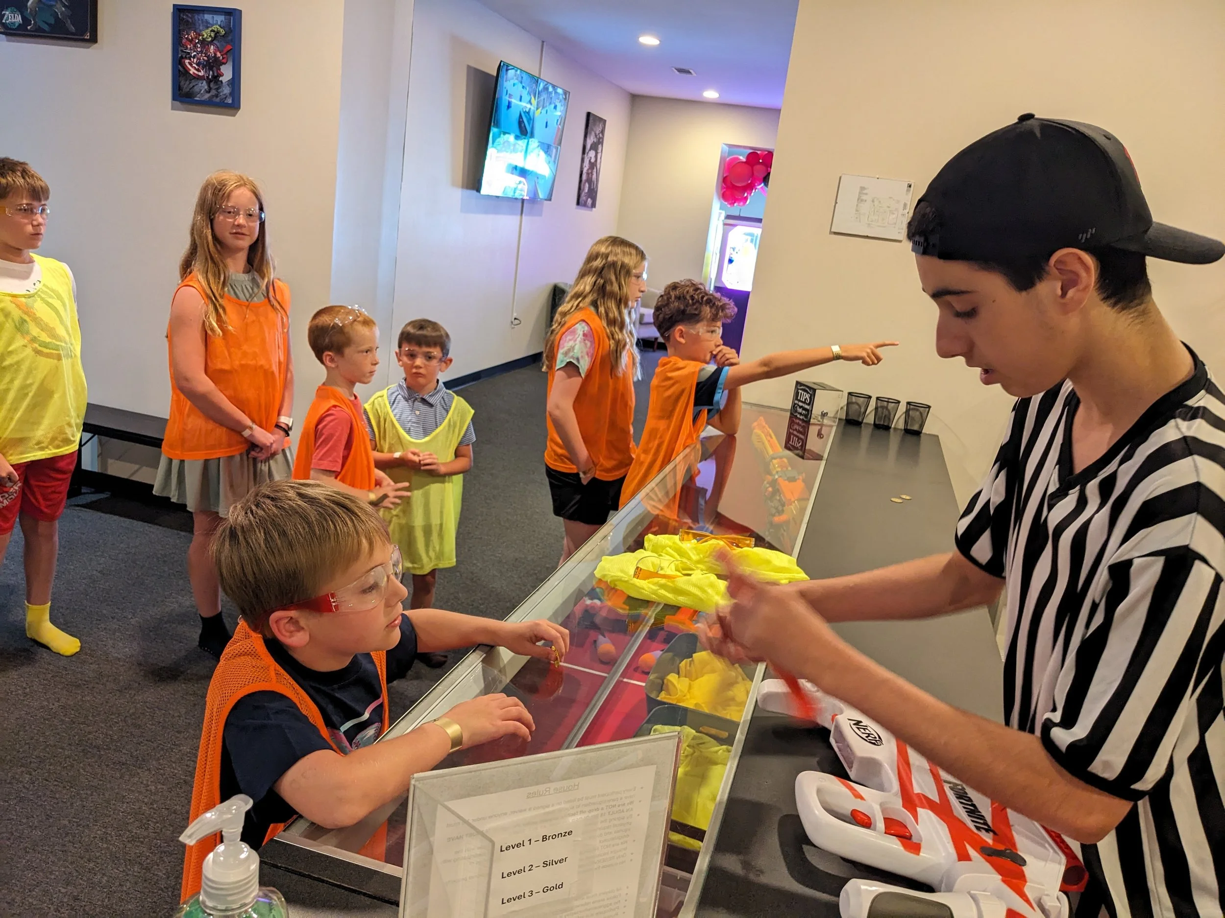 Children in line at an indoor arcade or activity center, with some wearing yellow vests and orange vests, and an employee in a striped shirt handing out yellow paper hats or bags.