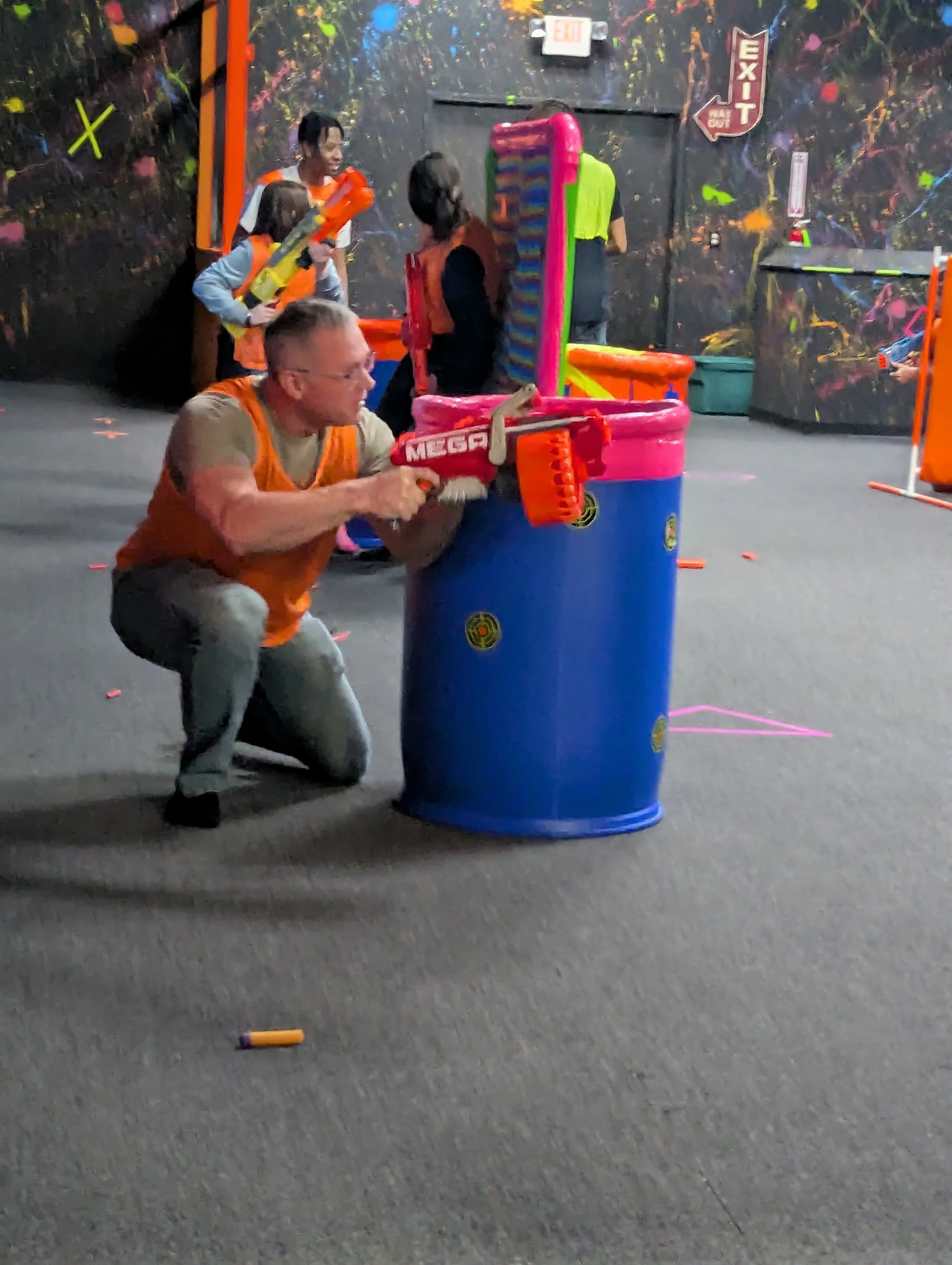 People playing Nerf battle in an indoor arena with dark, colorful, graffiti-style walls, some holding Nerf blasters, and one person kneeling behind a large blue barrel for cover.
