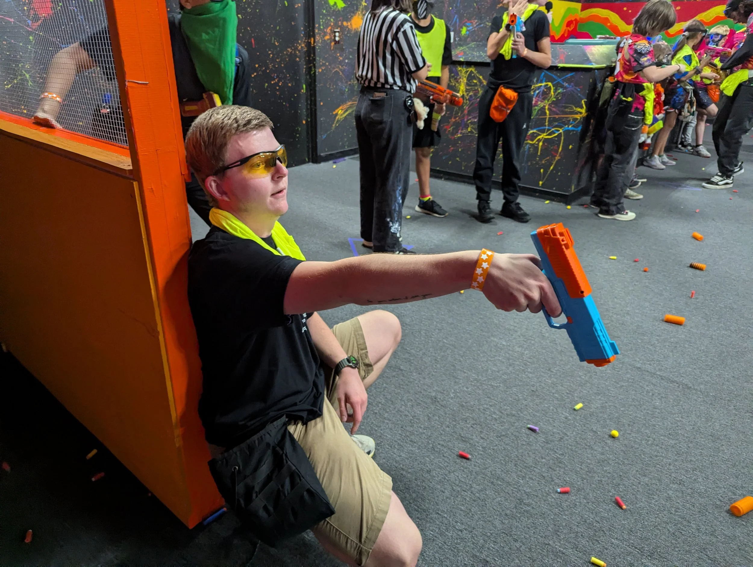 A young boy sitting on the floor behind an orange barrier, holding a Nerf gun with a yellow band on his wrist, wearing yellow-tinted safety glasses, black T-shirt, and khaki shorts. In the background, several people are standing and wearing safety gl