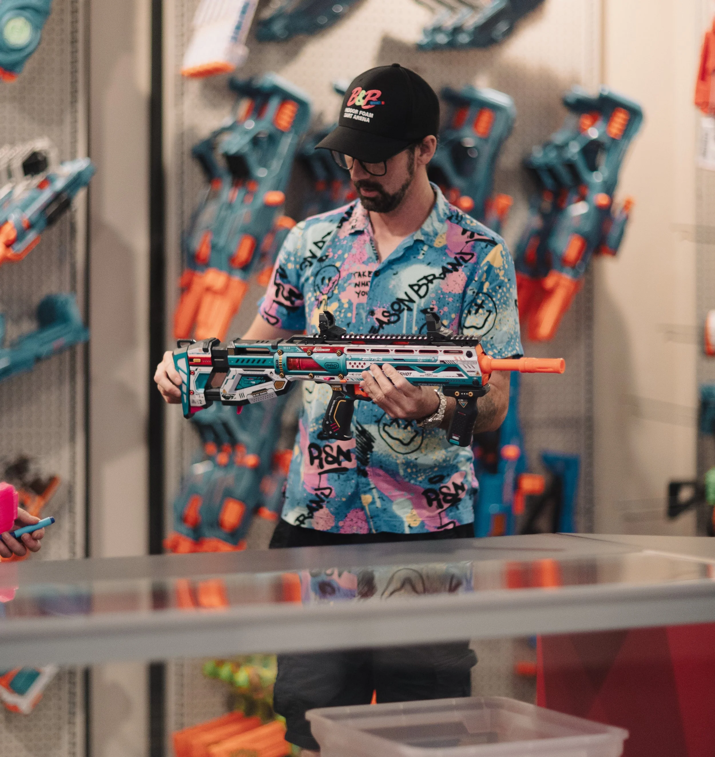 A man wearing a colorful, graffiti-style shirt and a black cap stands in a store aisle browsing a nerf gun with a futuristic design. He is holding the nerf gun with both hands, looking at it closely.