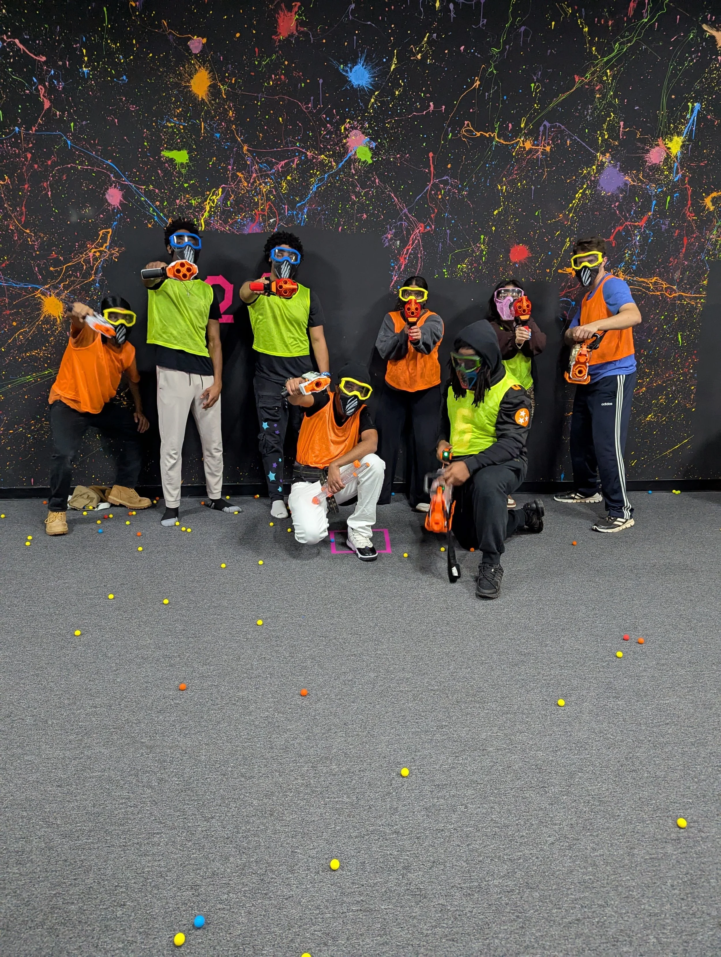 Group of people wearing safety goggles and holding foam dart blasters, posing in front of a colorful splatter pattern wall, with foam darts scattered on the floor.