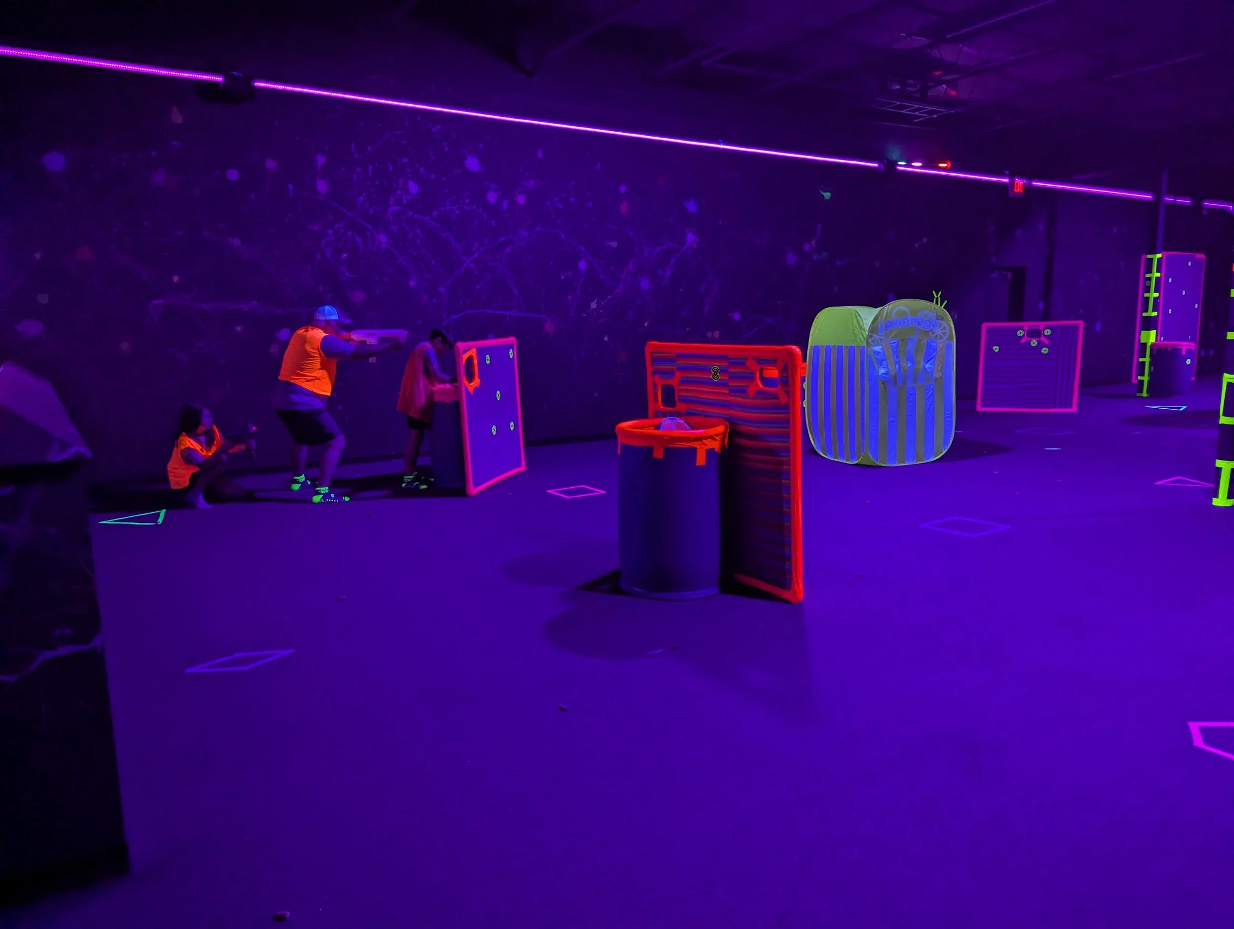 People playing laser tag in a dark room with neon lighting, glow sticks, and inflatable obstacles.