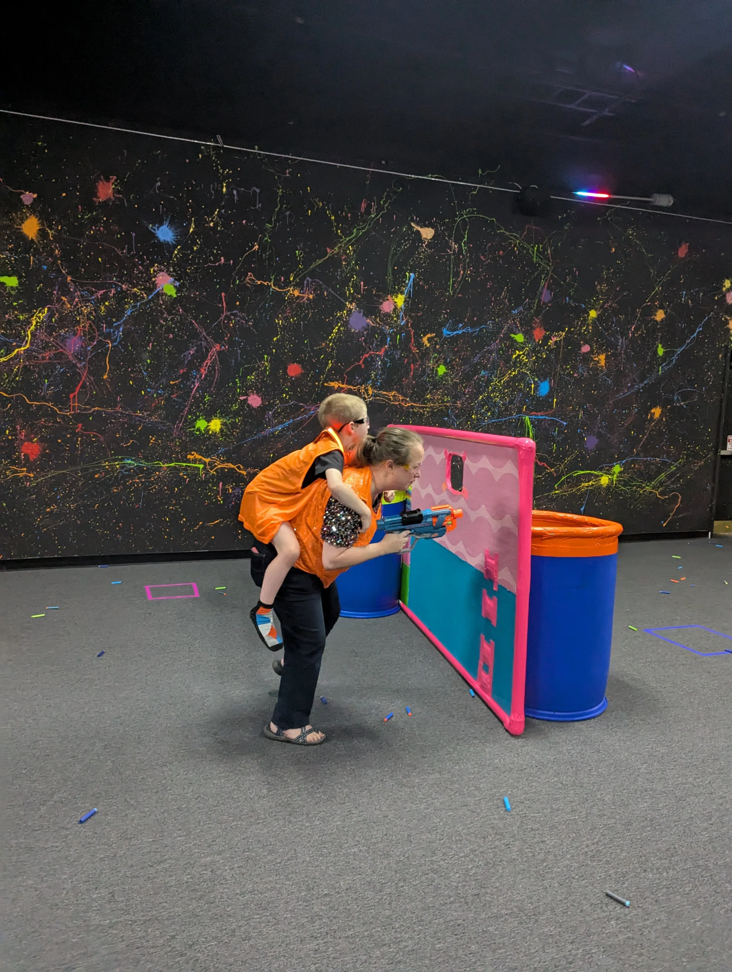 A woman giving a child a piggyback ride while playing with a toy gun behind a pink and blue plastic barrier at an indoor play area with black walls decorated with colorful paint splatters.