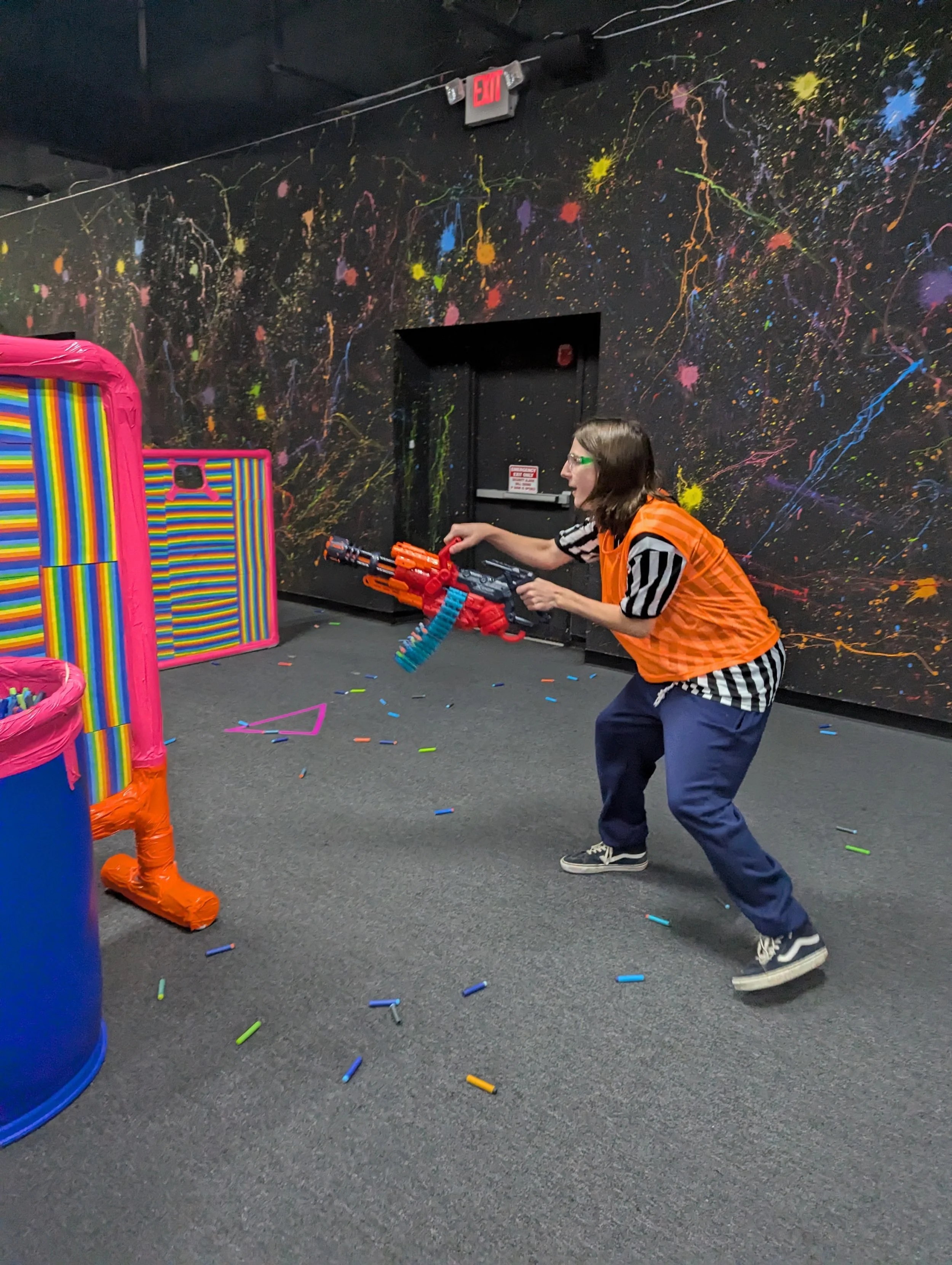 Woman in an orange vest playing with a Nerf blaster in an indoor laser tag or Nerf game arena with colorful confetti and spray paint splatters on the black walls.