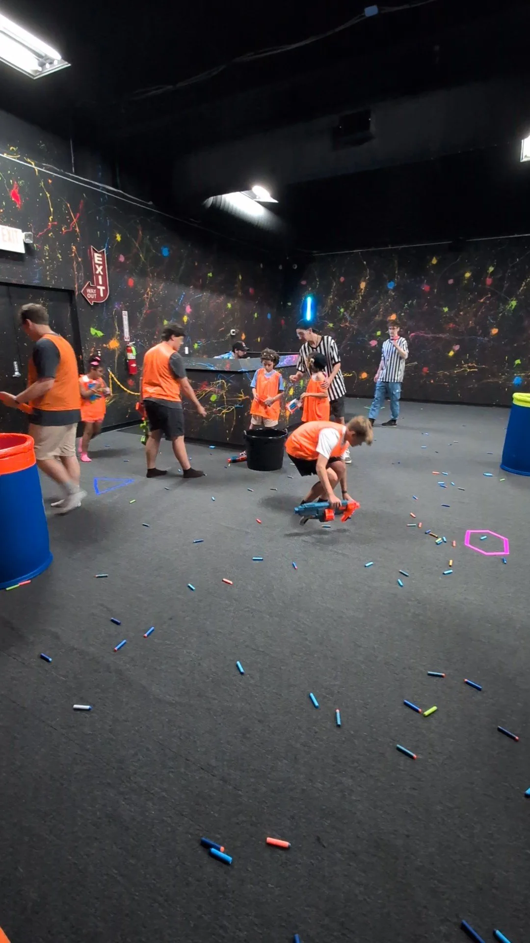 Children playing laser tag in a dark room with colorful wall splatters, neon lights, and scattered foam darts on the floor.