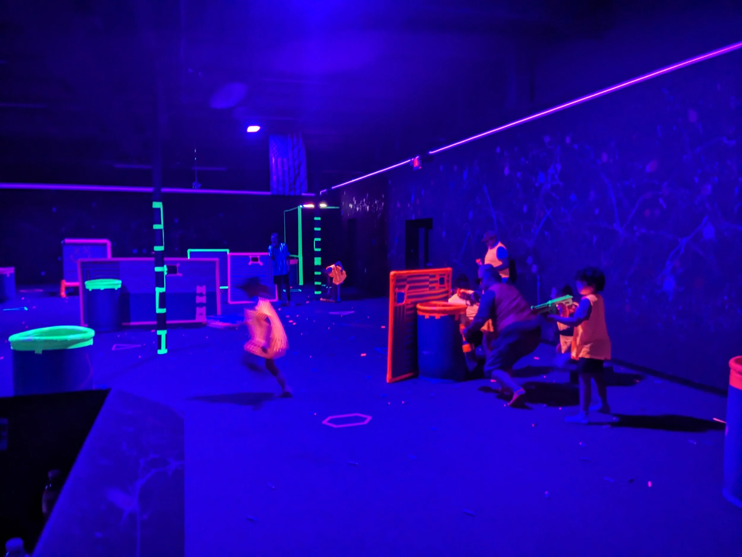 Children and adults playing laser tag in a dark room with neon lights and glowing obstacles.
