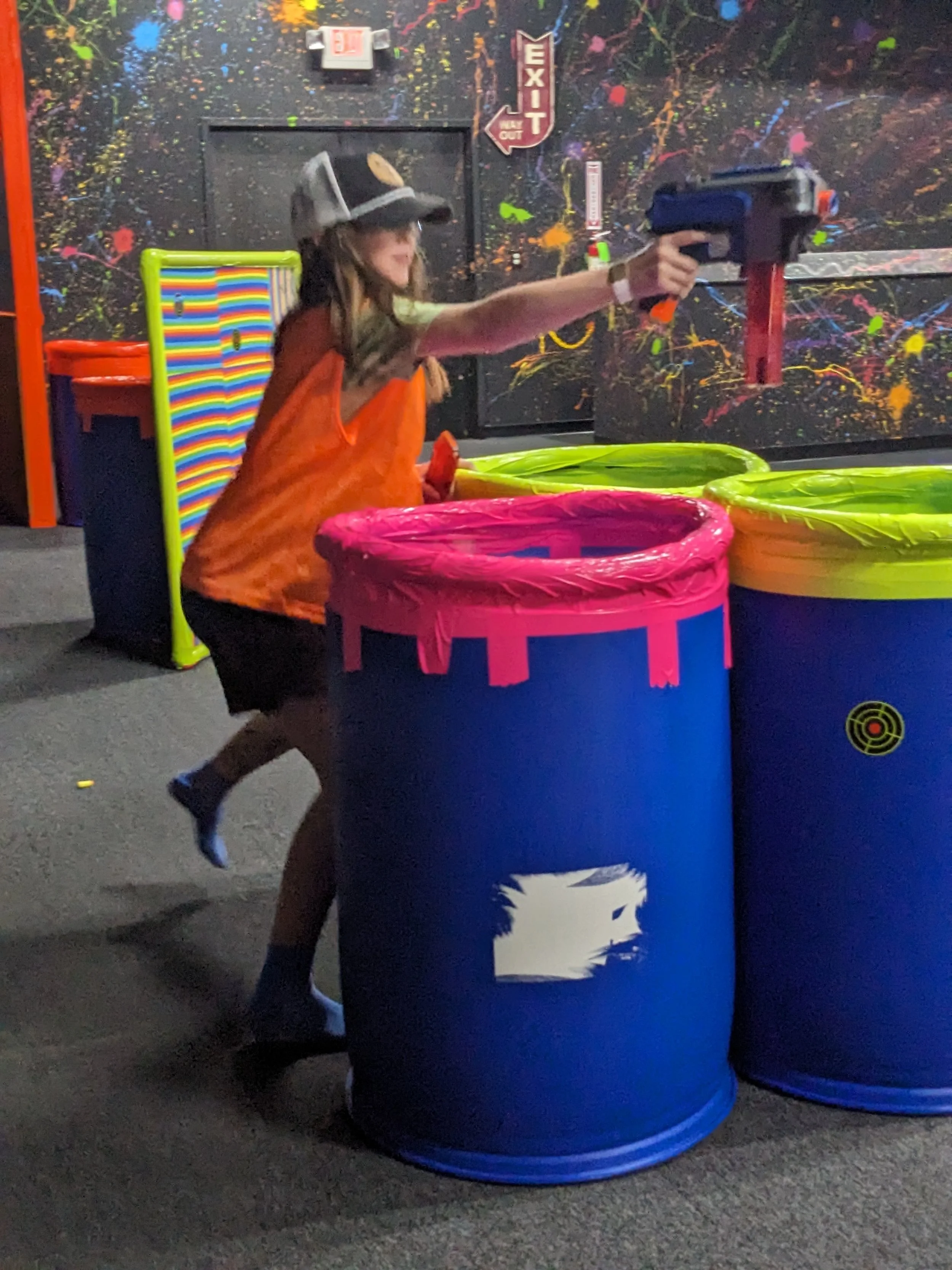 Child in an orange vest and a baseball cap playing nerf with a toy gun in an arcade with colorful barrels and graffiti-like wall.