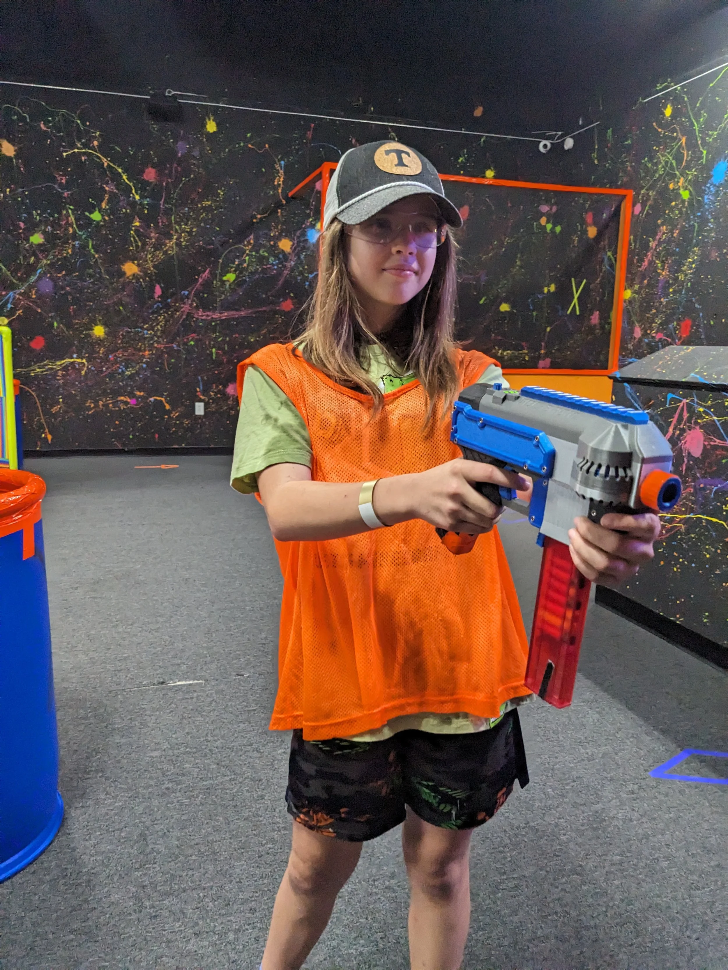 A girl wearing a gray baseball cap, safety glasses, an orange vest, a tan shirt, and camouflage shorts holding a colorful toy gun inside an indoor nerf arena with dark walls painted with colorful splatters.