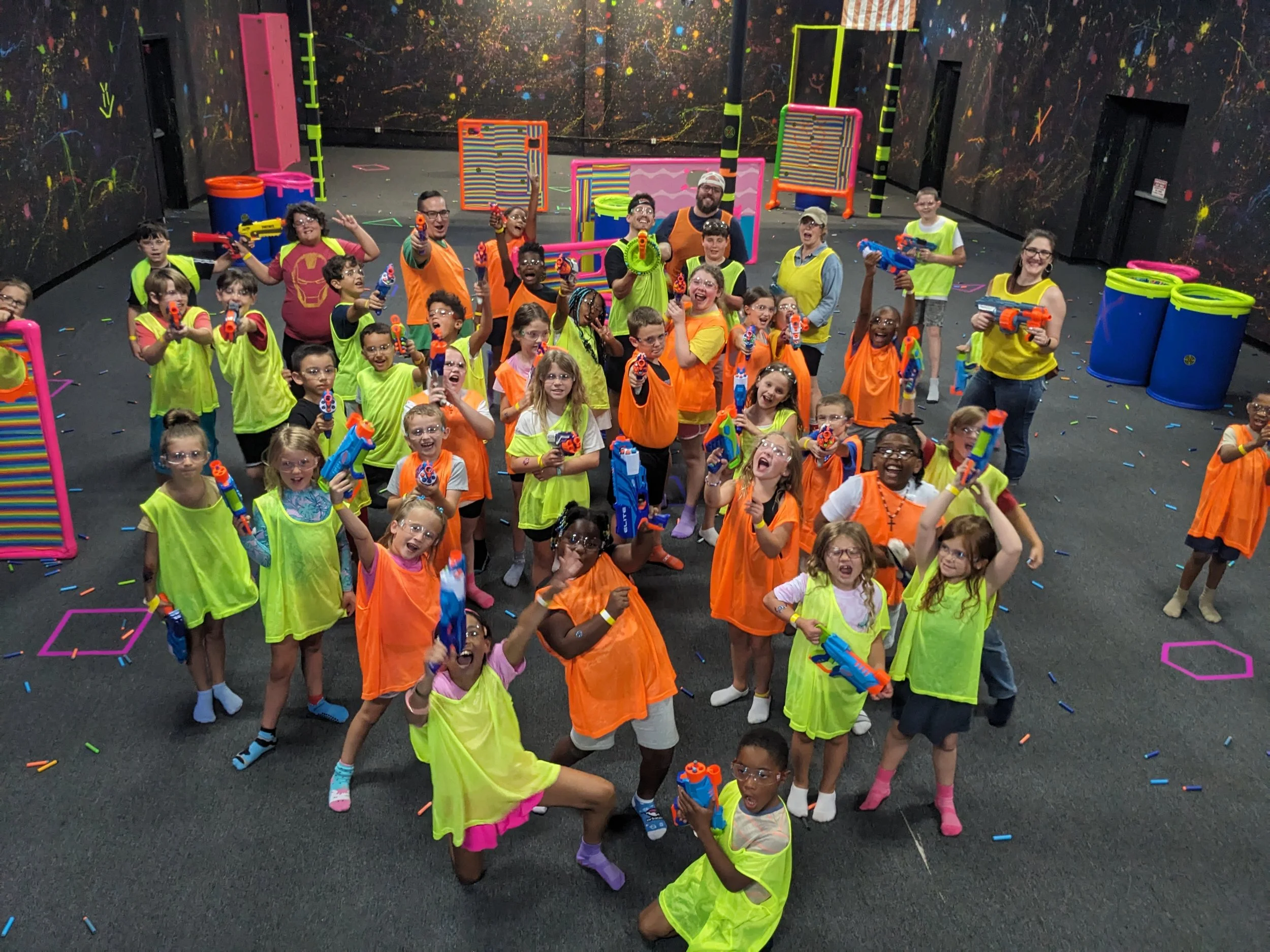 Group of children and adults playing laser tag indoors, wearing colored vests and holding toy laser guns, with vibrant decor and confetti on the floor.