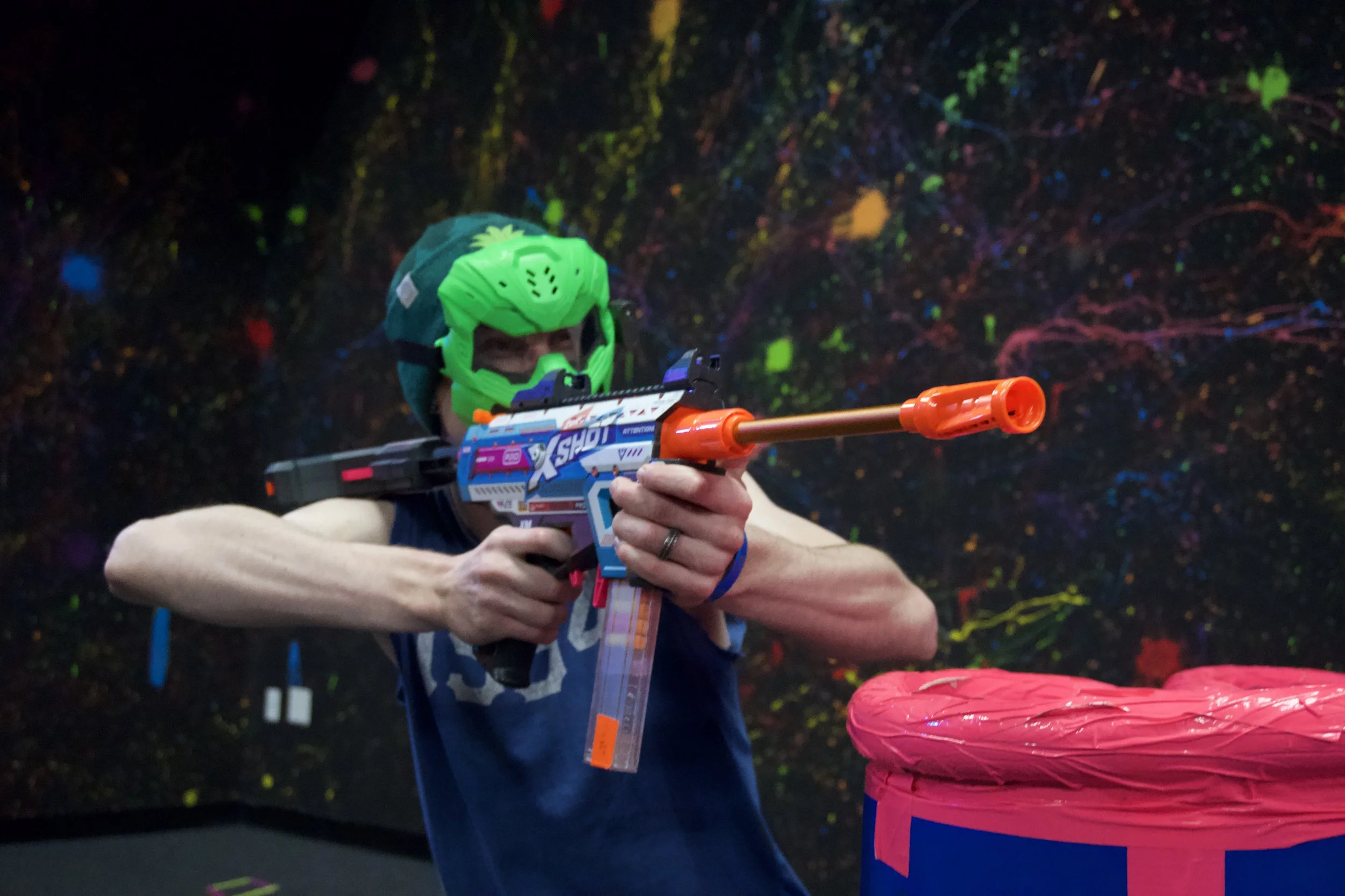 Person wearing a green helmet aiming a Nerf blaster, with a neon colorful background.
