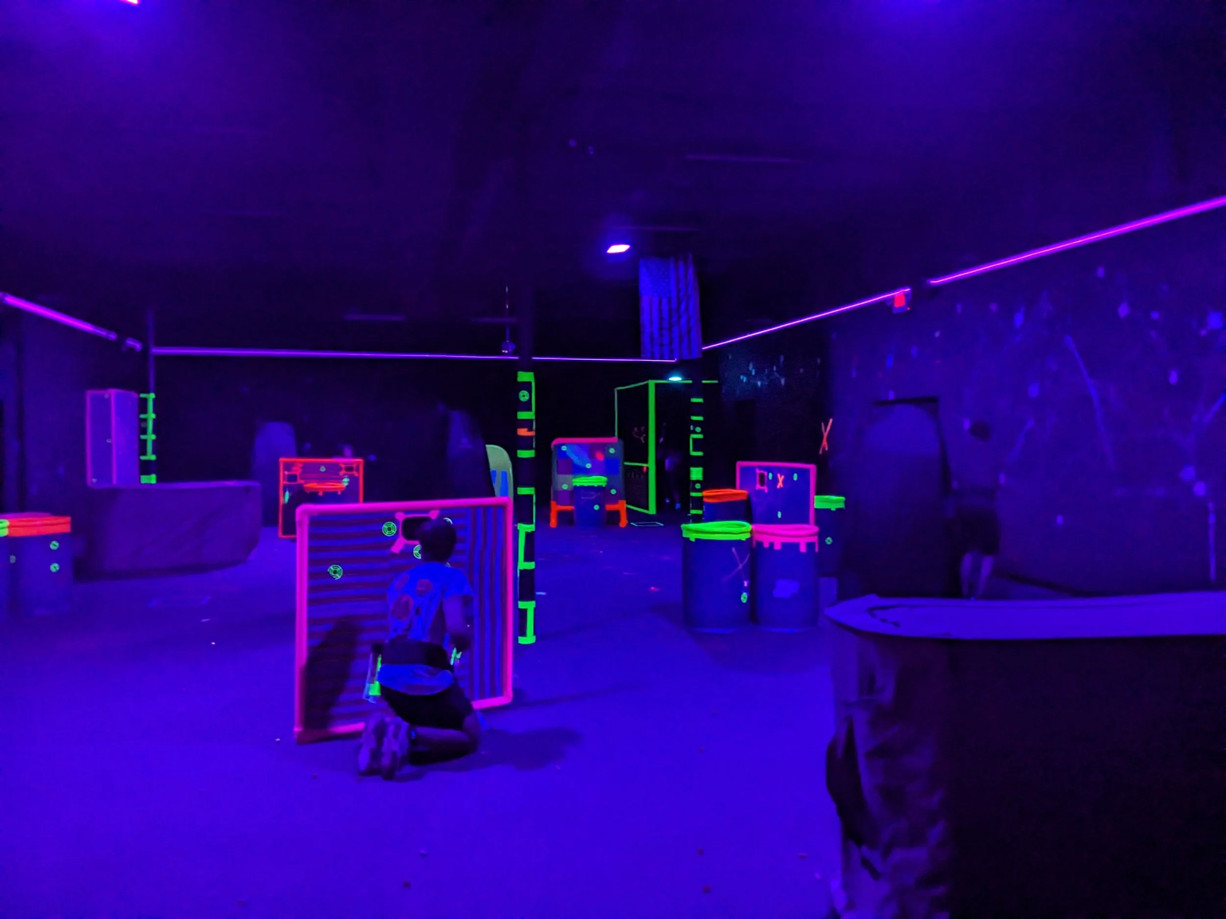 Dark indoor laser tag arena with neon-lit barriers and walls, kids playing with laser guns under blacklight.