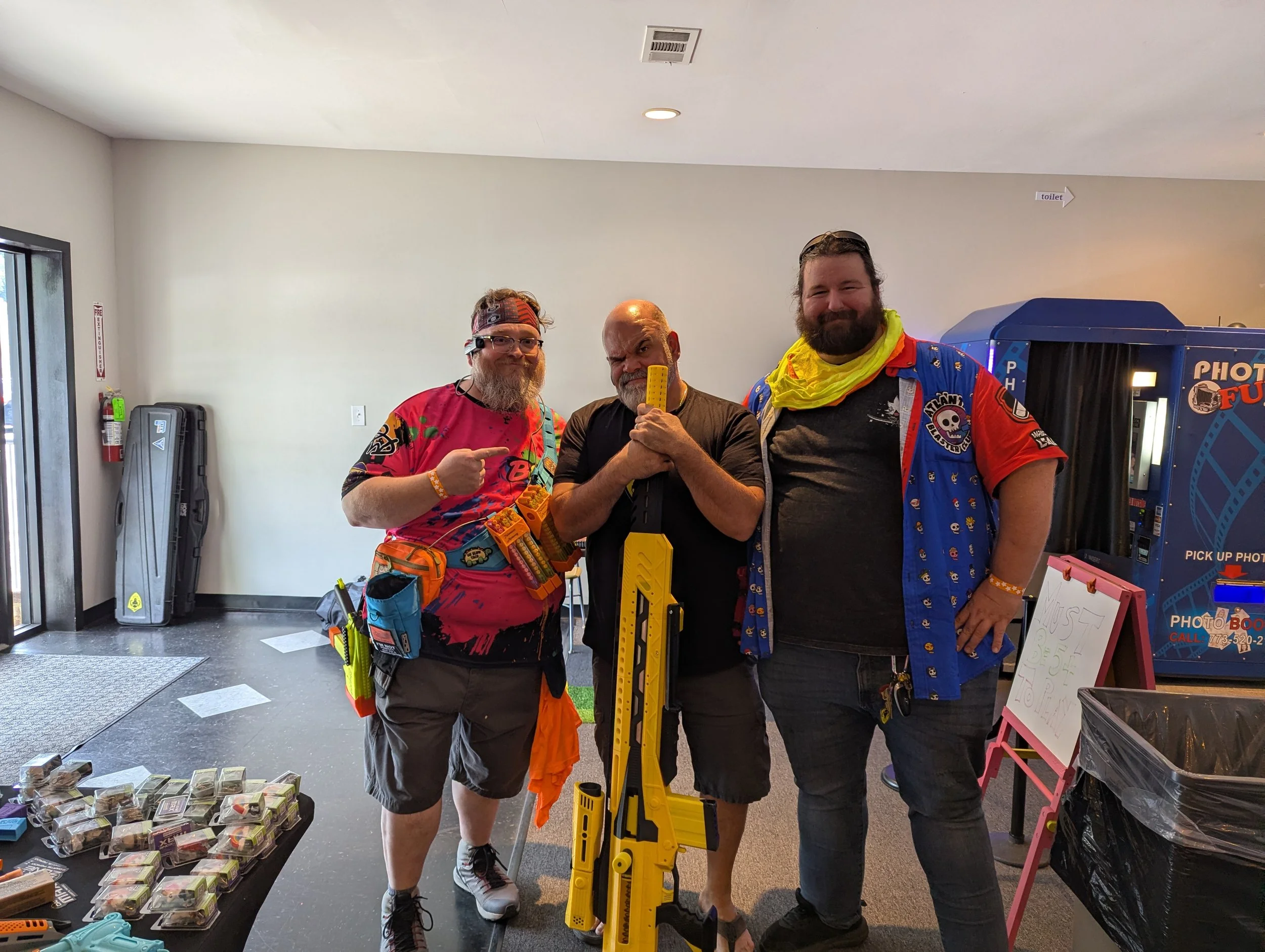 Three men standing indoors, smiling and posing for the camera. The man on the left has a beard, glasses, and is wearing a colorful shirt and a headband. The man in the middle, who is bald with a gray beard, is holding a yellow tape measure. The man o